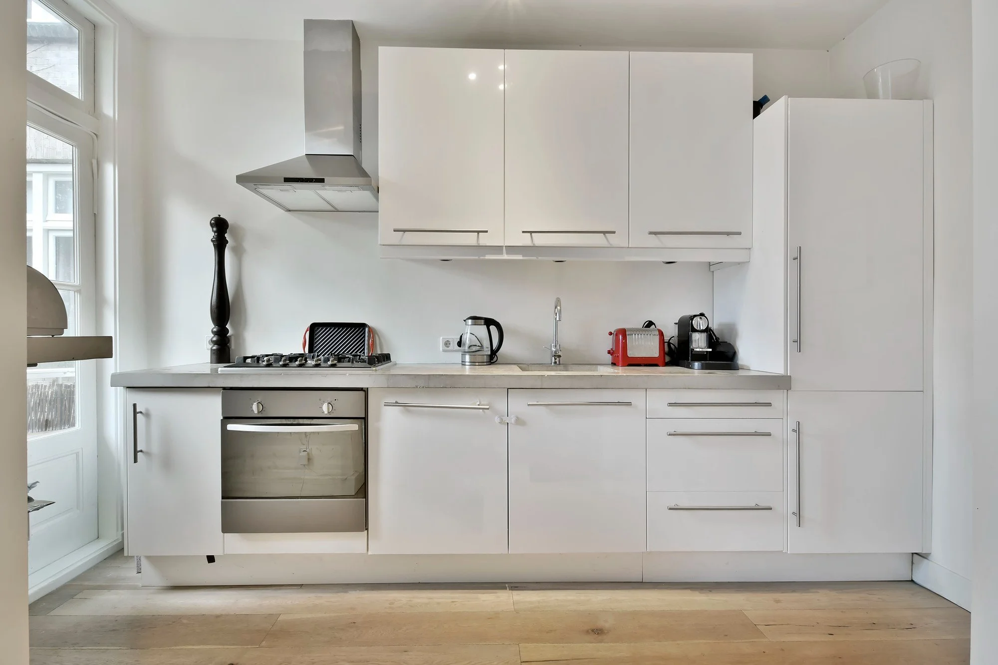 Modern white kitchen with stainless steel appliances, including an oven and a range hood, and small appliances on the counter such as a kettle, toaster, and coffee machine. White cabinets above and below, with a hardwood floor.