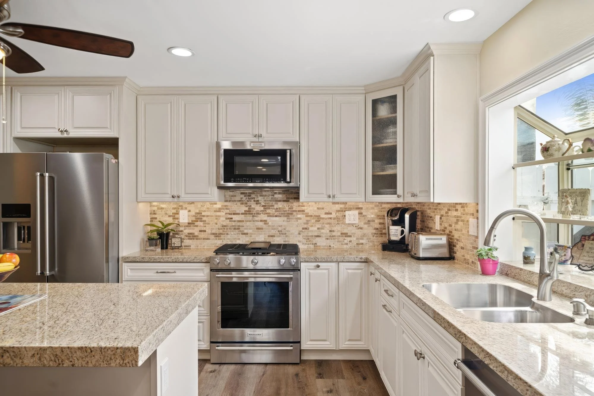 Modern kitchen with white cabinetry, stainless steel appliances, granite countertops, beige tile backsplash, and a large window with a pink potted plant on the sink
