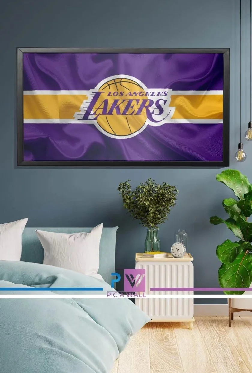 Living room with a framed Los Angeles Lakers basketball team flag or banner on the wall, a bed with white pillows, a white nightstand with a plant, a clock, and a glass container, and green plants in the room.