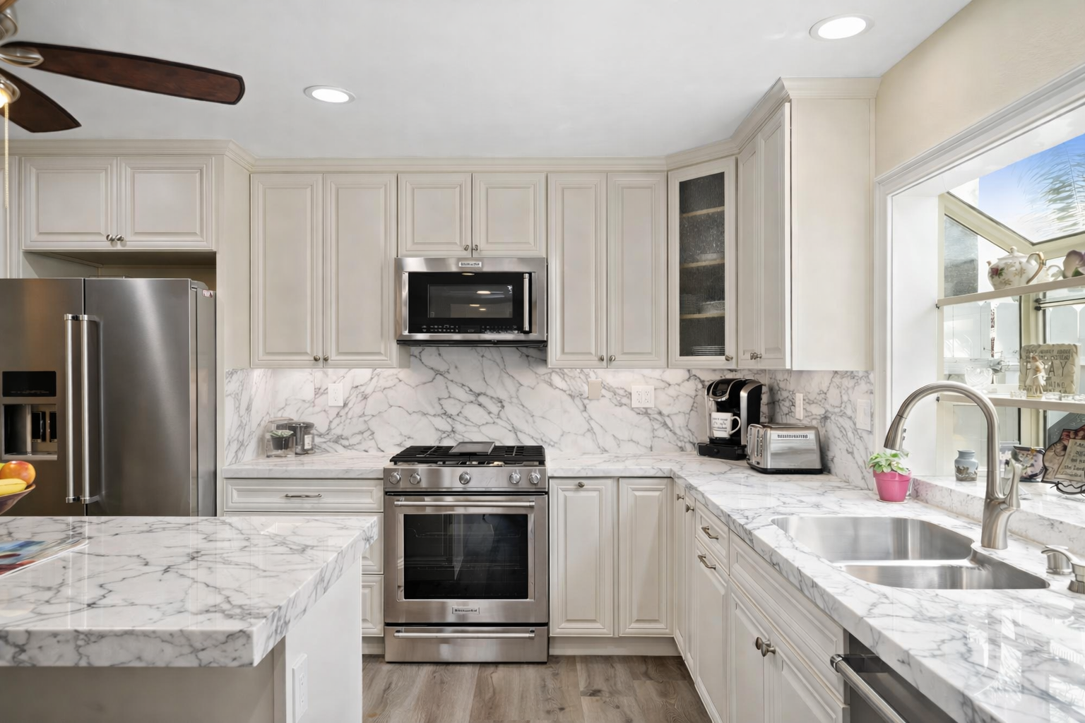 Modern kitchen with white cabinetry, marble countertops and backsplash, stainless steel appliances, and a window above the sink.