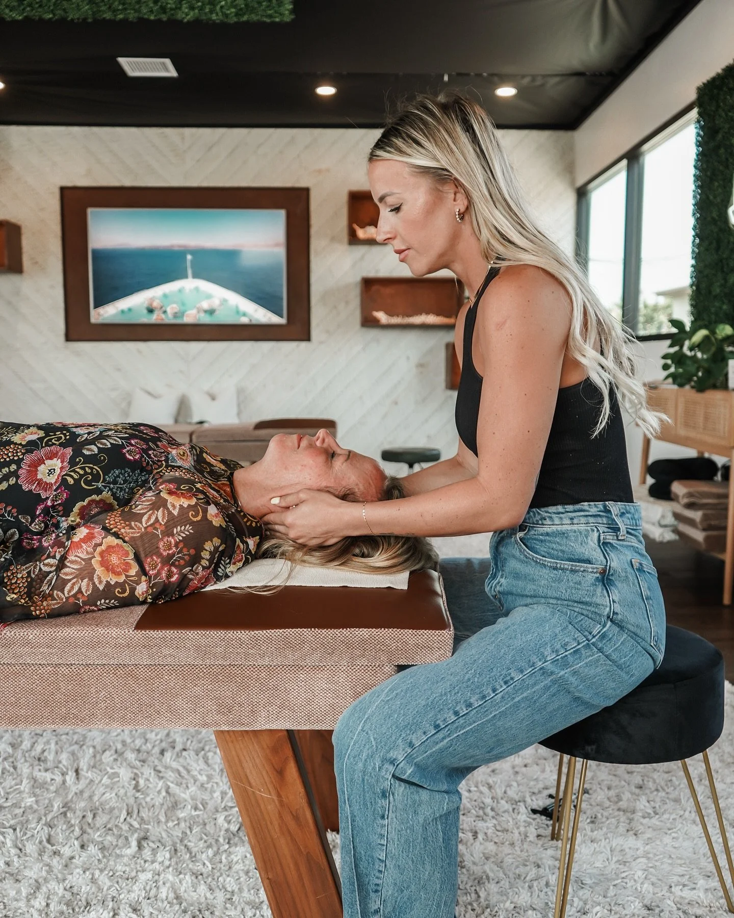 Woman with long blonde hair providing a facial massage to an elderly woman on a massage table in a well-lit room with modern decor.