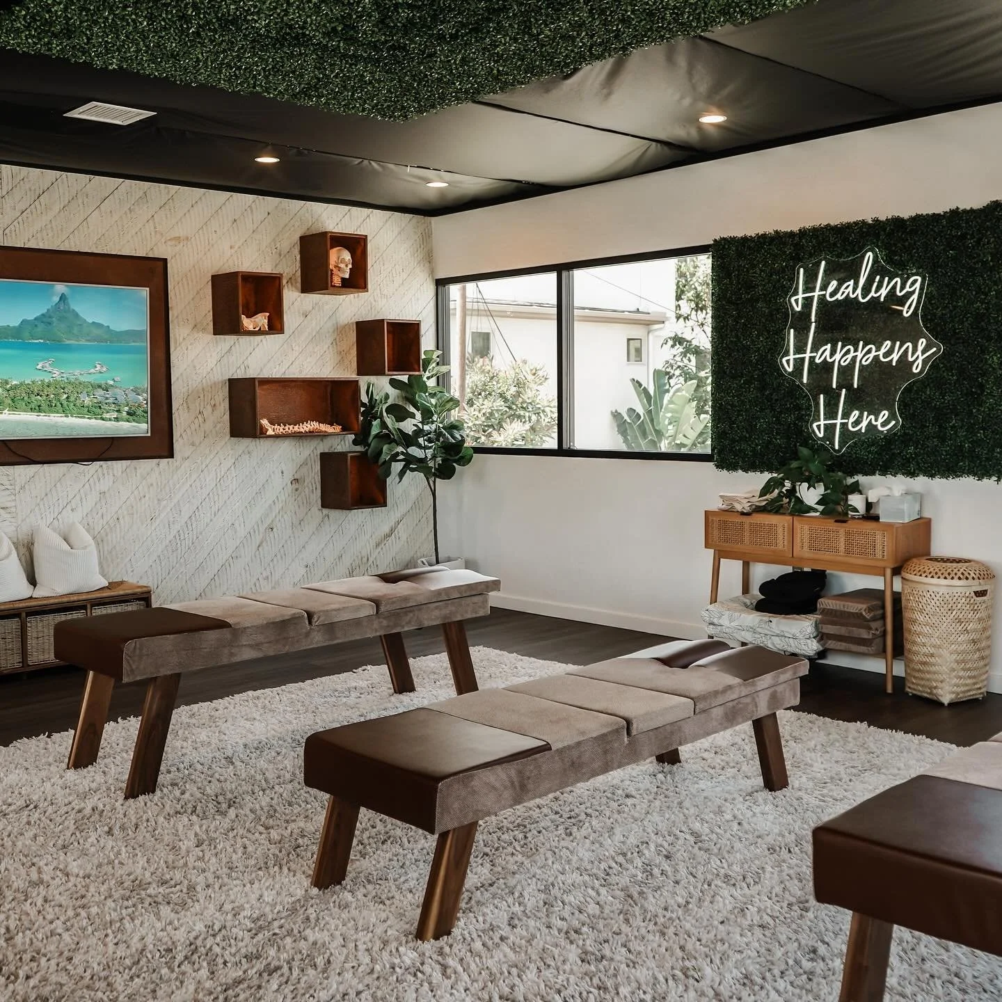 A wellness or therapy room with benches on a plush white rug, wall-mounted shelves with decorative objects, a wall-mounted TV, large window, potted plant, and a green wall with neon sign reading