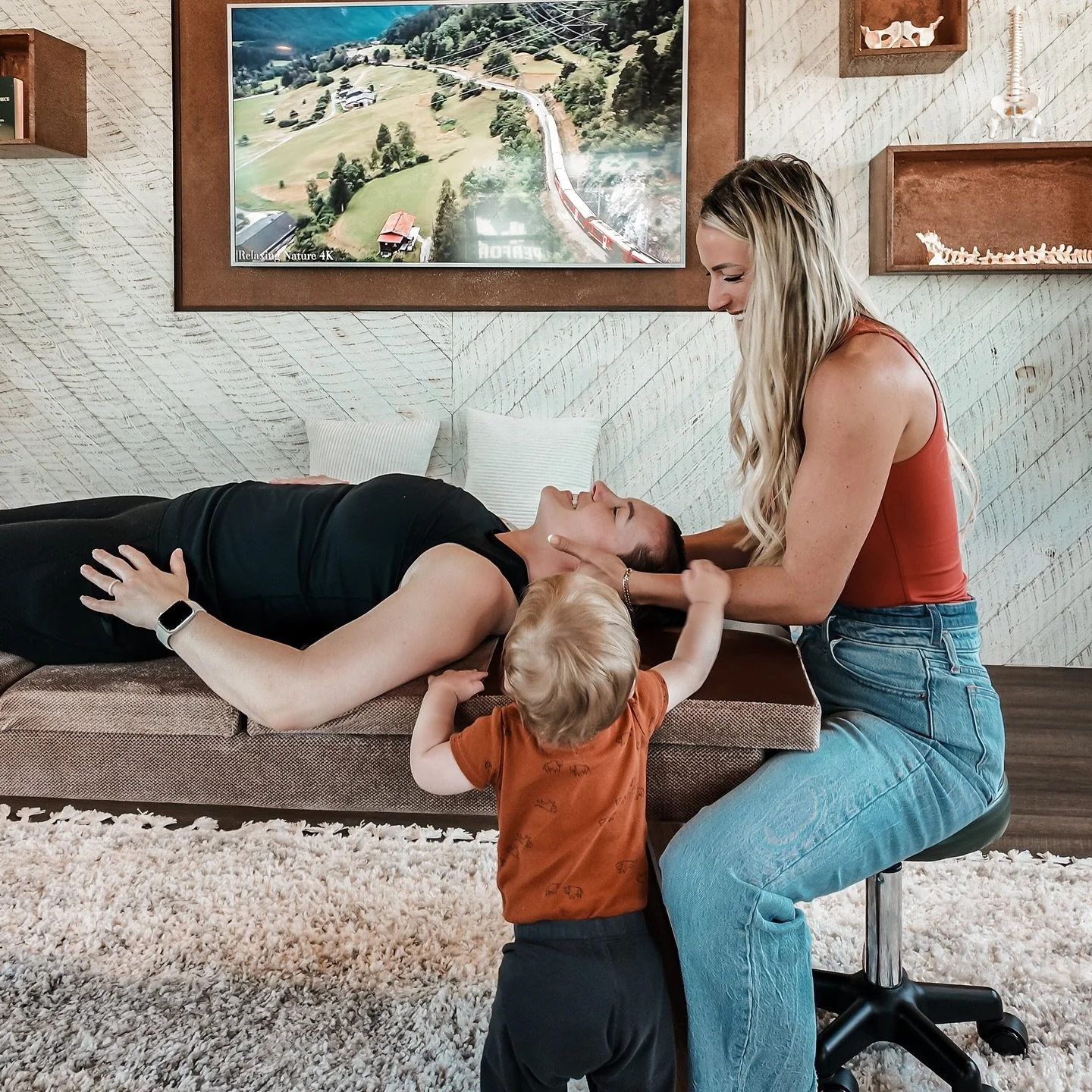 A woman lying on a couch with her eyes closed, while another woman with long blonde hair is performing a check on her neck. A young child with blonde hair, wearing an orange shirt, is standing and touching the woman’s face. They are in a cozy living room with a TV on the wall and decorative items.