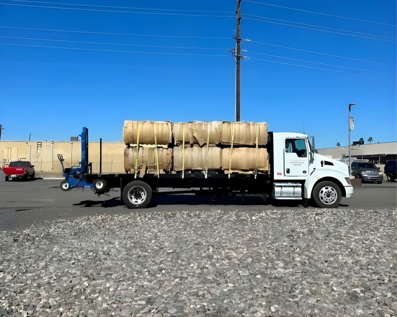 A white flatbed truck parked on a gravel lot, carrying beige cylindrical tanks secured with yellow straps. Several other vehicles and buildings are visible in the background under a clear blue sky.