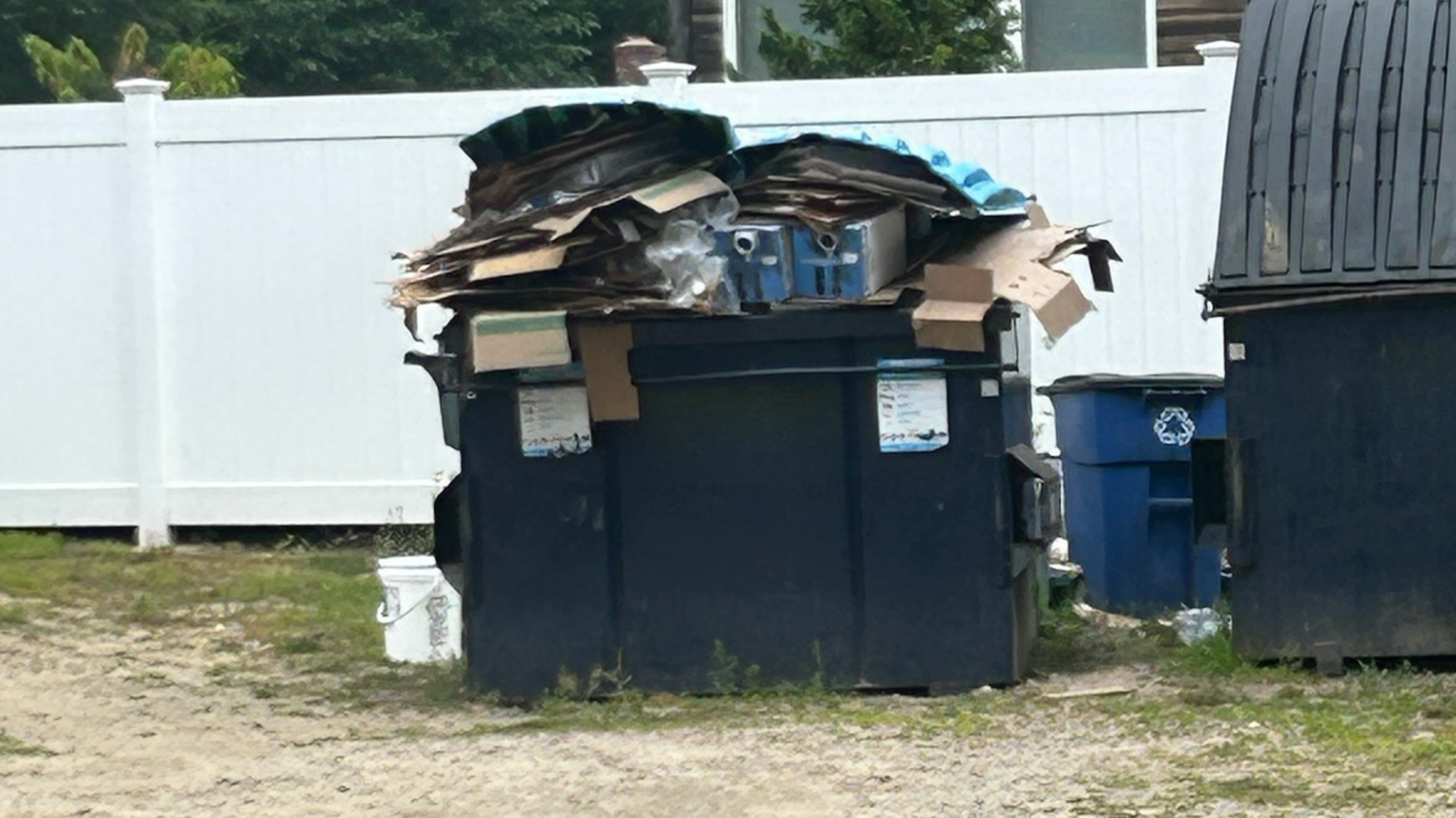 A large black trash dumpster filled with flattened cardboard and other waste, with a white privacy fence in the background and another smaller blue trash bin nearby.