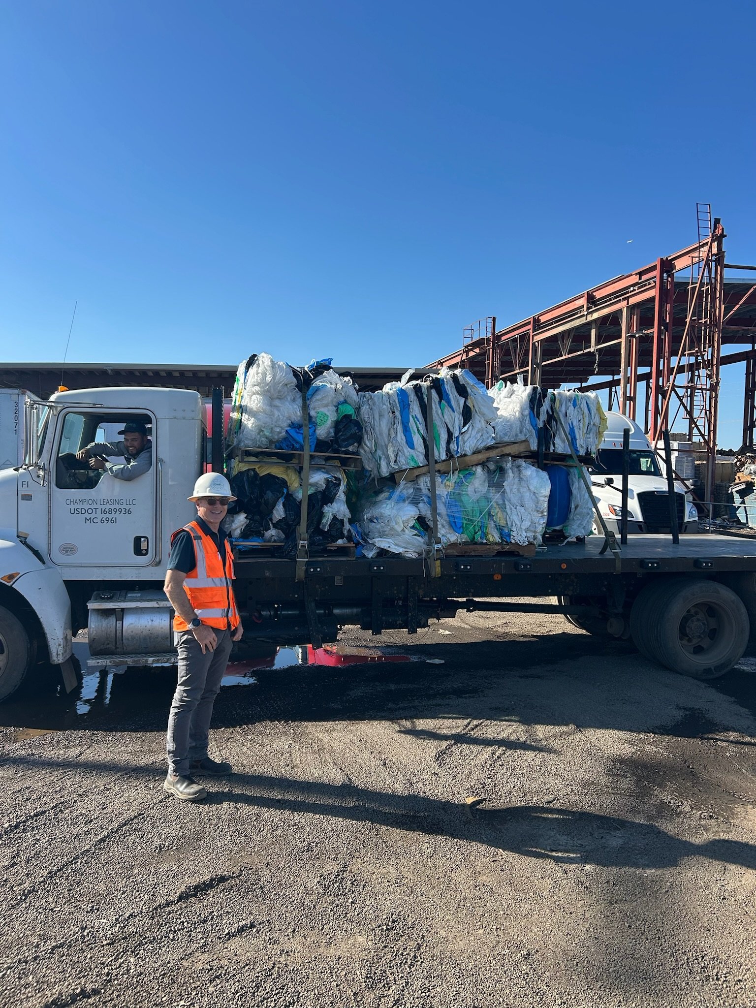 A man and a woman in safety gear posing in front of a large truck loaded with compressed recyclables at a construction or recycling site.