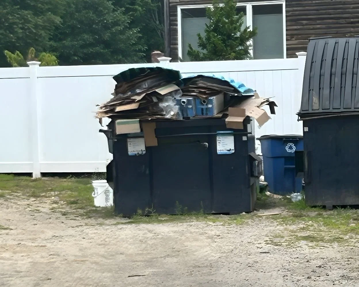 An overflowing dumpster with cardboard boxes piled high, next to recycling bins and a white fence in a residential area.