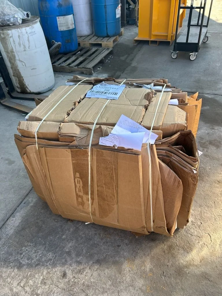 A large bundle of damaged, flattened cardboard boxes secured with white plastic straps, sits on a concrete surface in an industrial or warehouse setting.