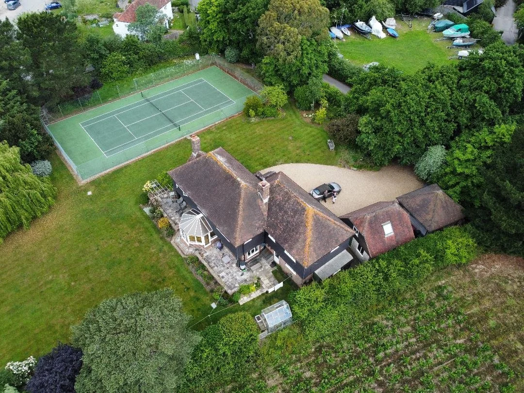 An aerial view of a large backyard with a tennis court, a house with a patio, and a gravel driveway with a car parked.