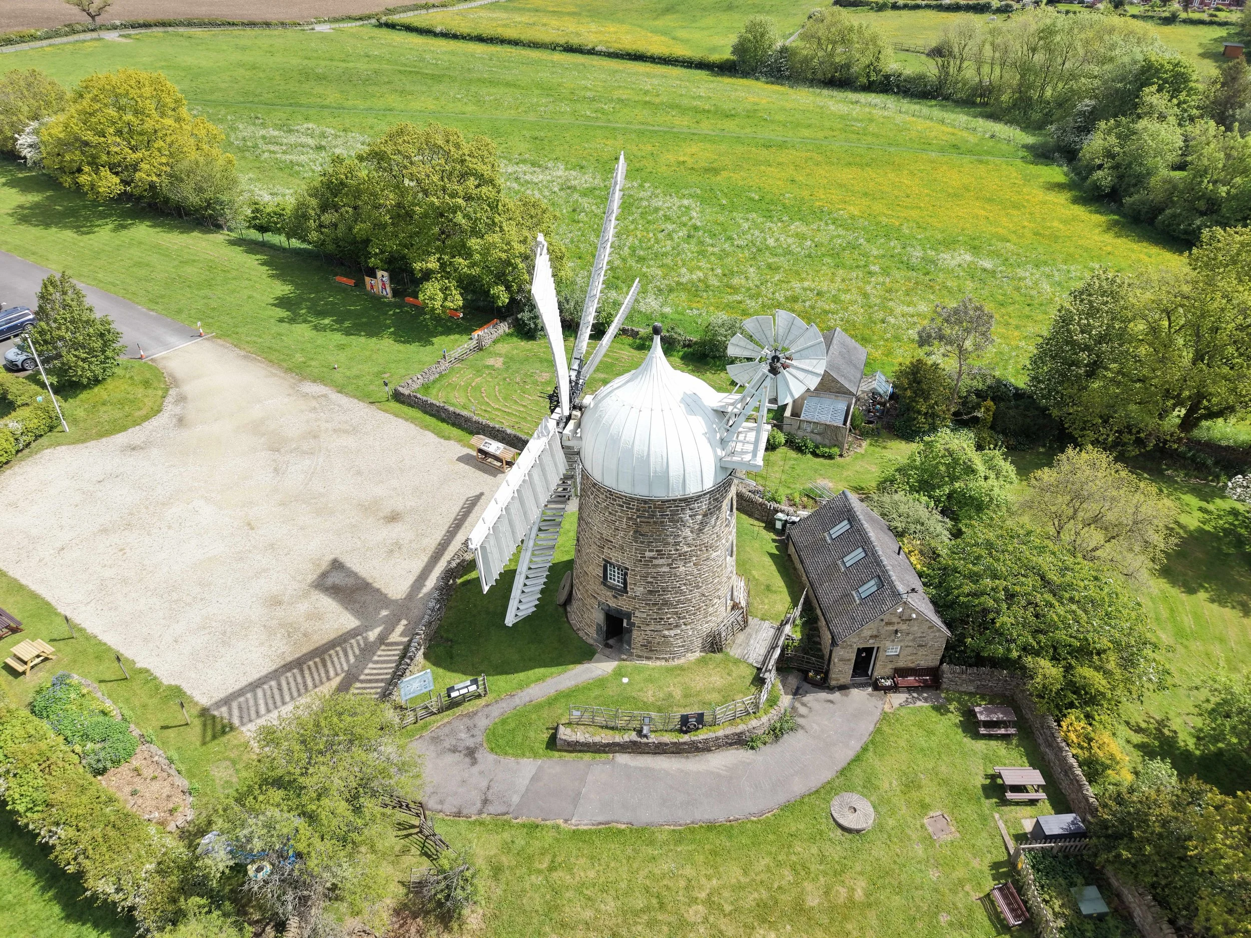 An aerial view of a historic windmill with white blades and a stone tower, situated in a green park with trees, grass, and walking paths, near a parking lot and some small houses.