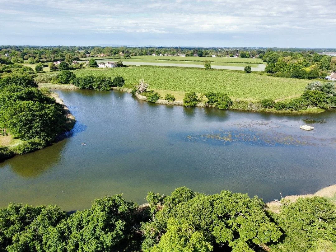 Aerial view of a lake surrounded by green trees, fields, and scattered houses under a partly cloudy sky.