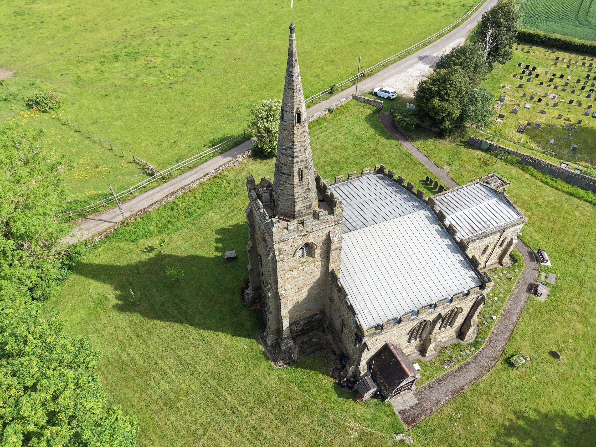 Aerial view of an old stone church with tall steeple, adjacent cemetery, green grassy area, trees, a small road, and a white car parked nearby.