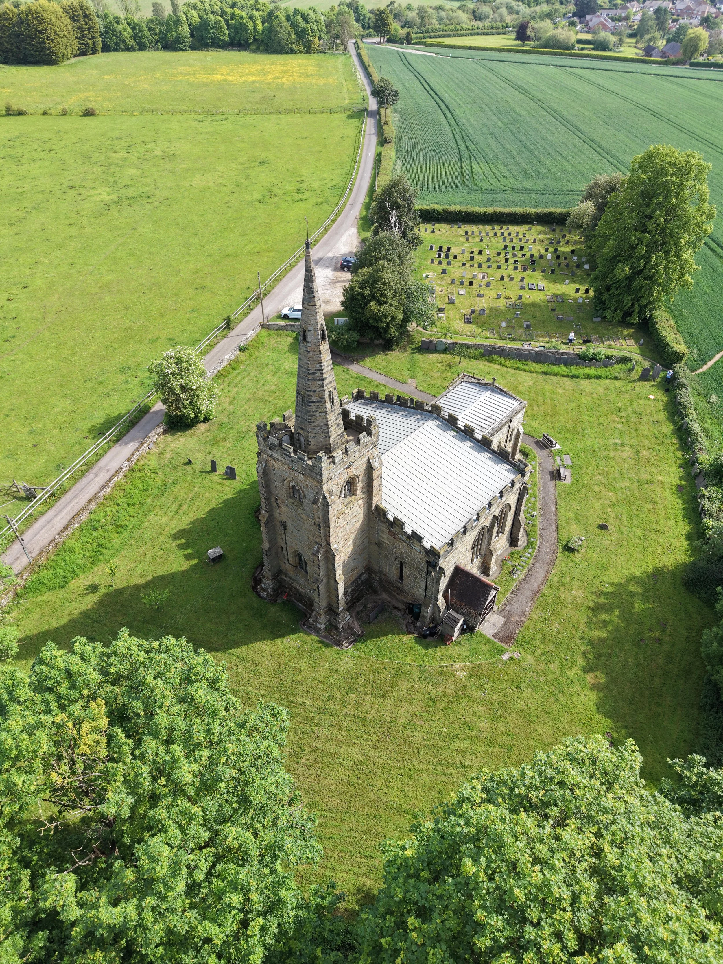 Aerial view of a church with a tall, pointed steeple, surrounded by green trees and a graveyard, with fields and a small town in the background.