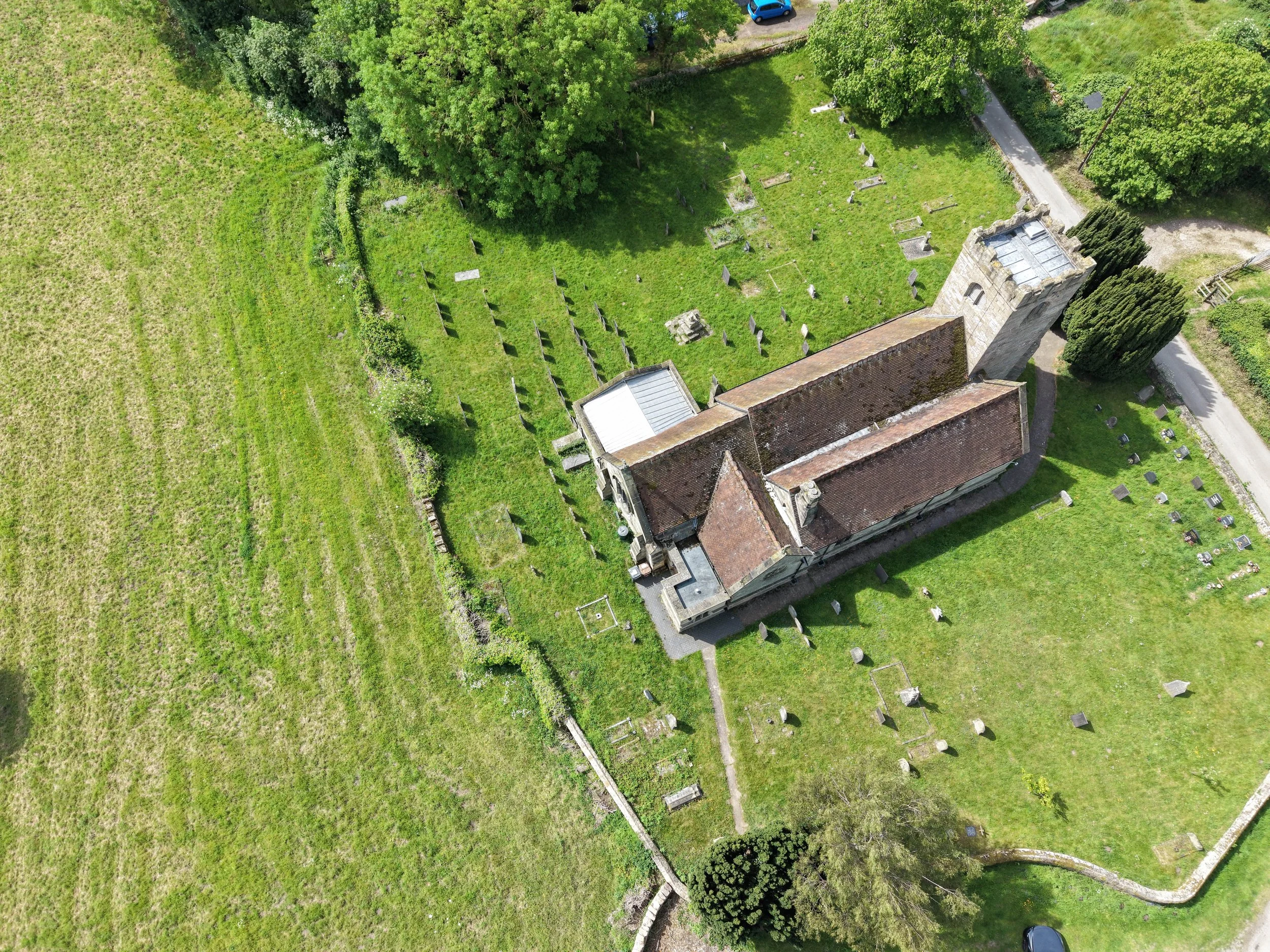 An aerial view of a church yard with tombstones and a church building with a red roof, trees, and a small gravel pathway. There are cars parked on a road beside the church yard.