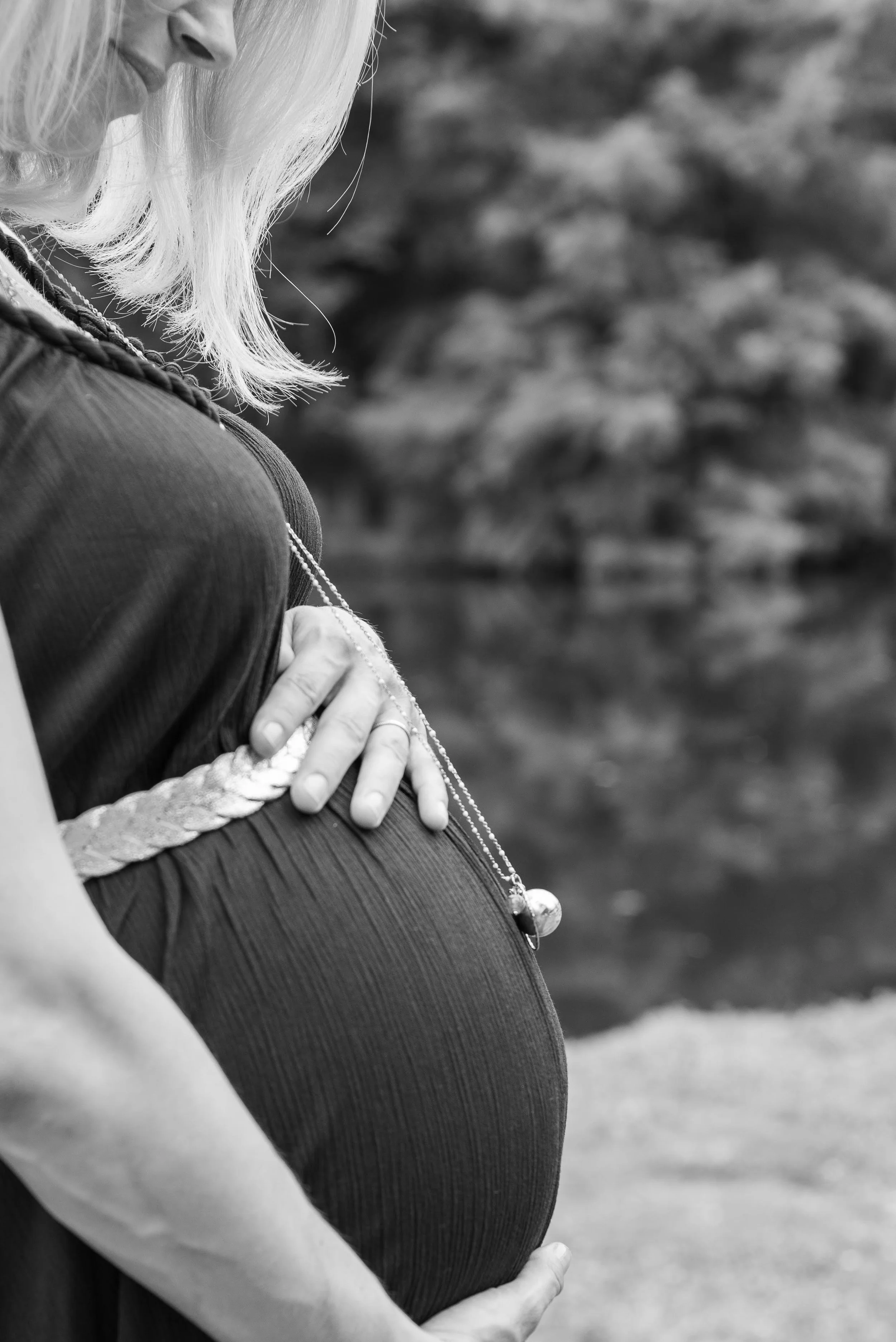 Femme enceinte tenant son ventre arrondi, en noir et blanc, près d'un plan d'eau.