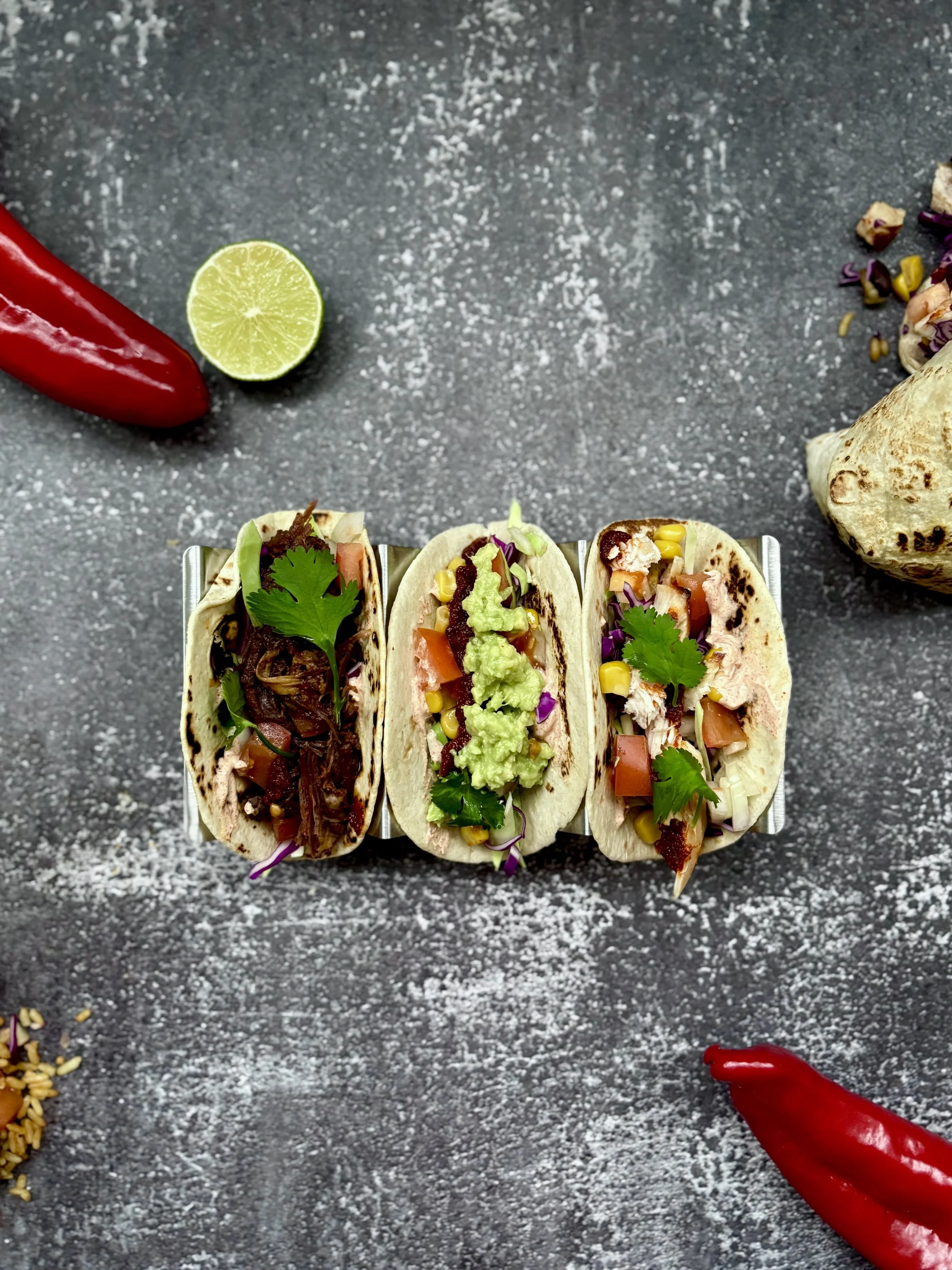 Tre Mexicanske tacos i Aarhus fyldt med kød, grøntsager, guacamole og koriander, lagt på en sort bord med lime og chili omkring.