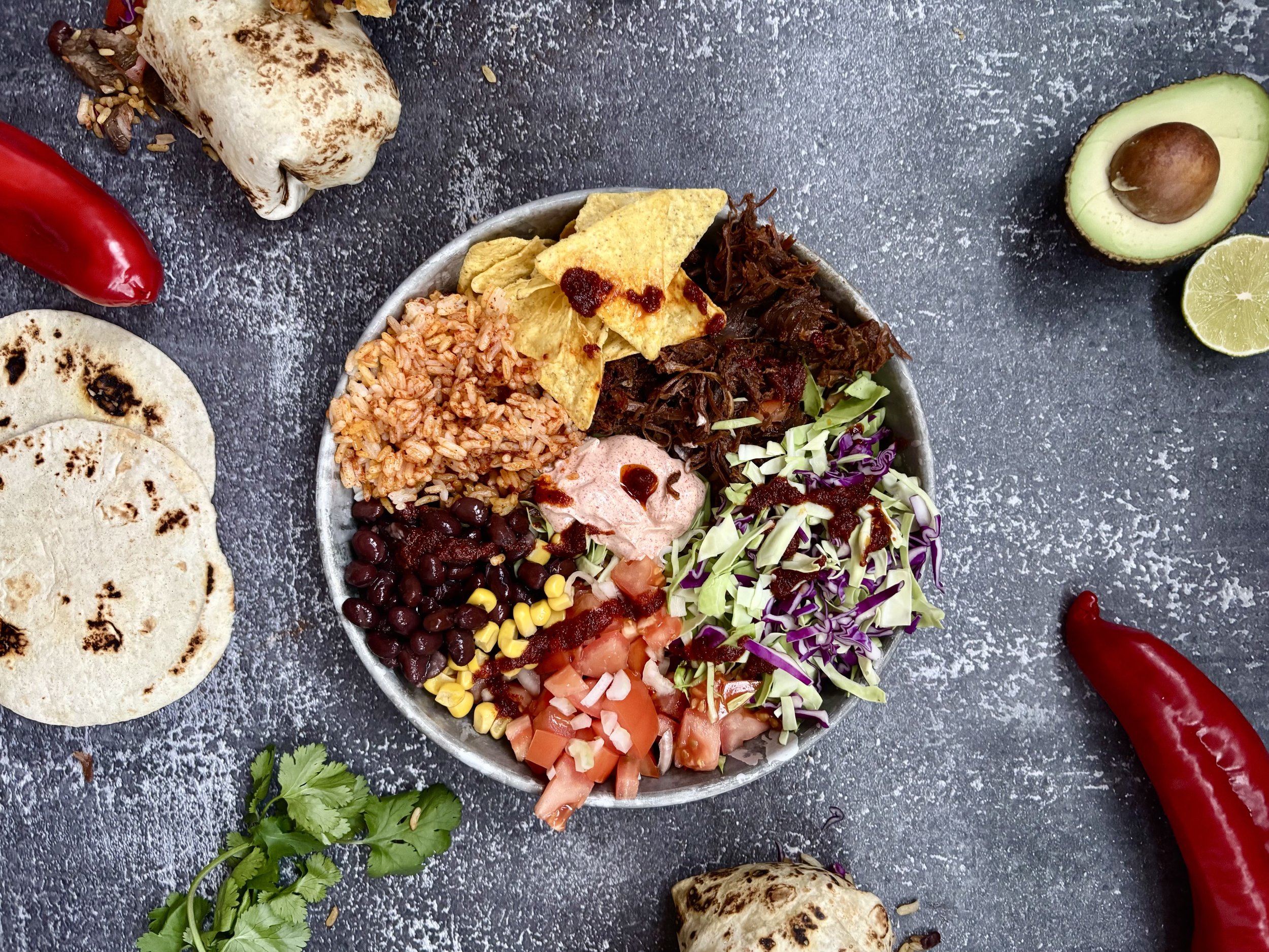 En skål med mexicansk mad i Aarhus, med ris, bønner, salat, tomater, avokado, ost, tortillachips, grøntsager og sauce, omgivet af chili, lime, tortillapit, koriander og røgede grøntsager på en sort baggrund.