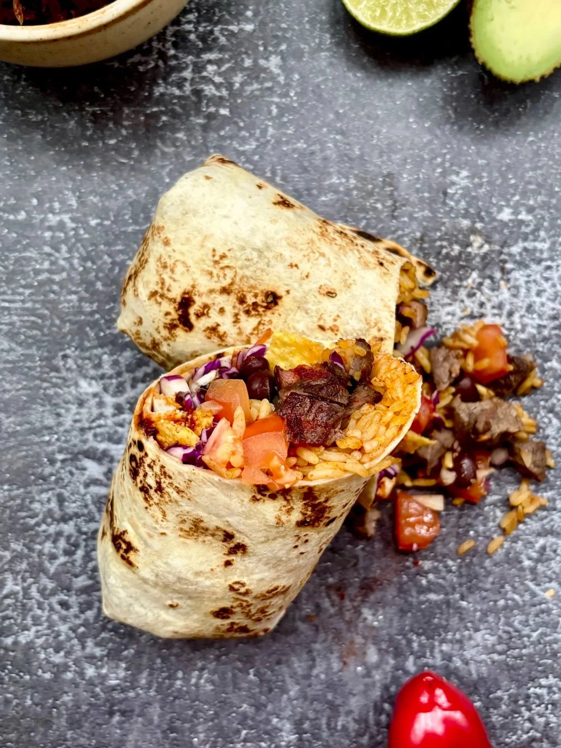Mexicansk burritos i Aarhus, fyldt med ris, kød, grøntsager og bønner, placeret på en mørk overflade med lime, cherrytomater og en skål i baggrunden.