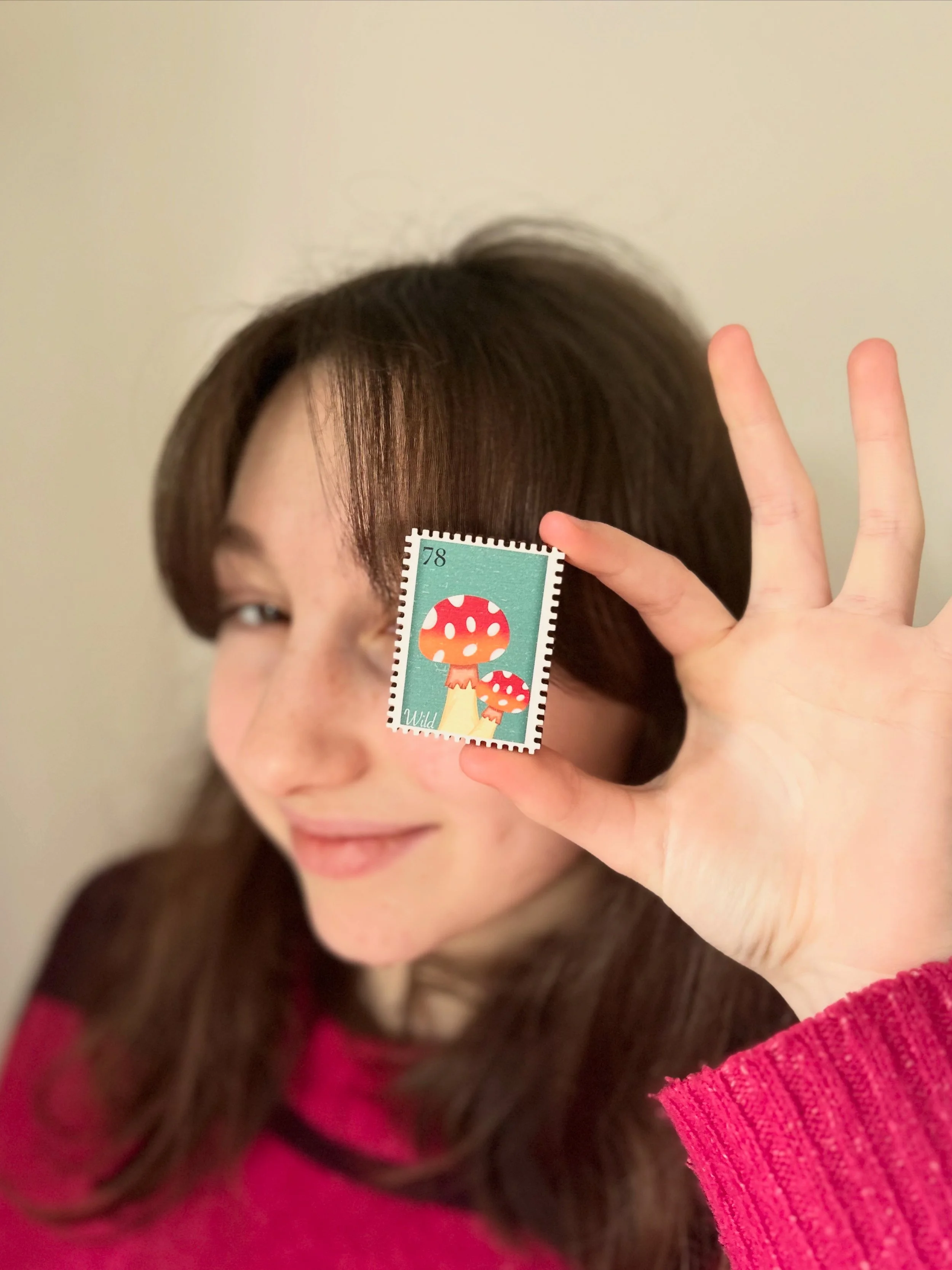 Girl with long brown hair holding up a stamp with colorful mushroom illustrations in front of her eye.