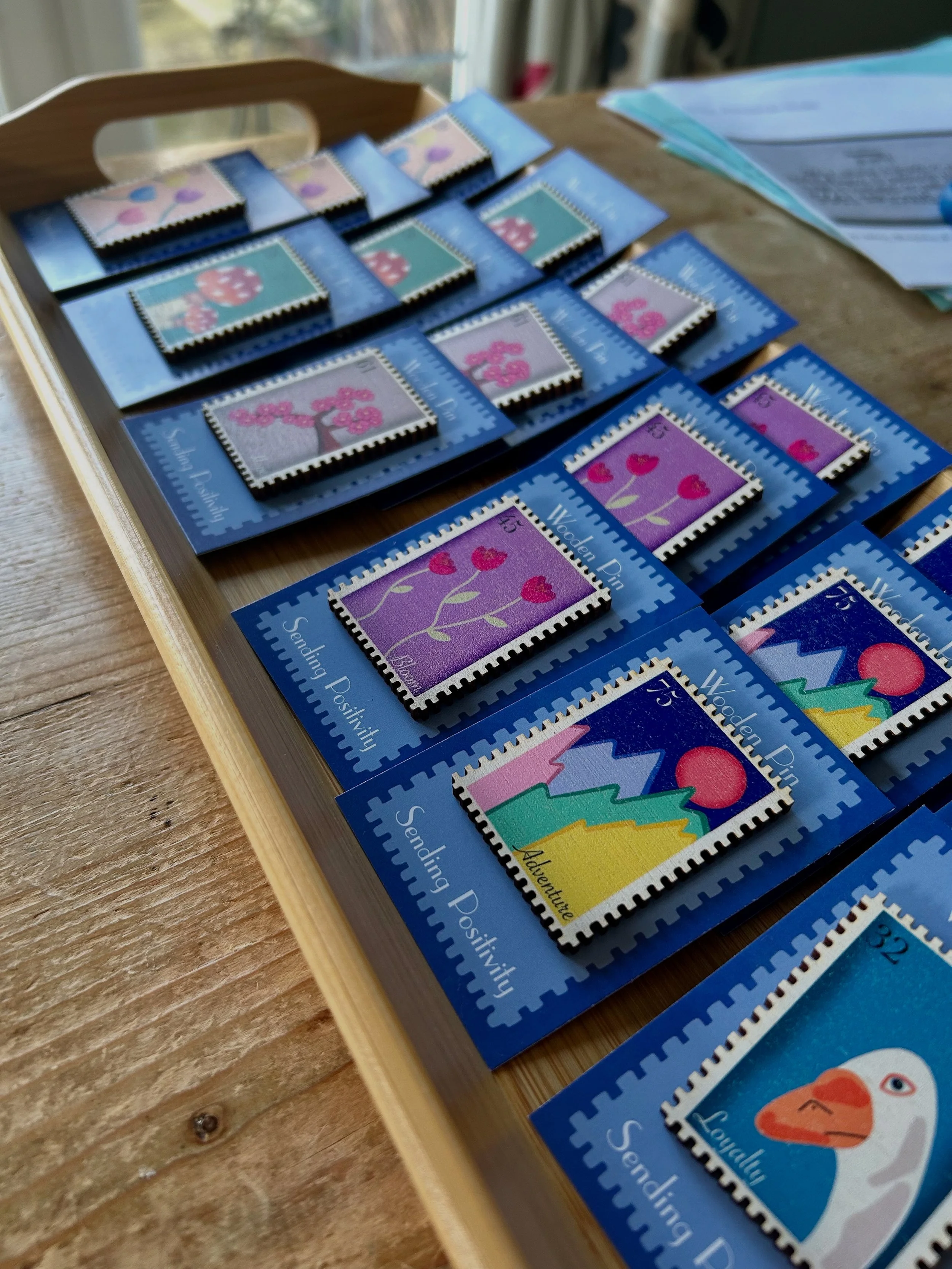 A wooden tray holding colorful illustrated stickers in clear plastic sleeves, each labeled with a theme like 'Adventure' or 'Sending Positivity,' on a wooden table with papers and documents nearby.