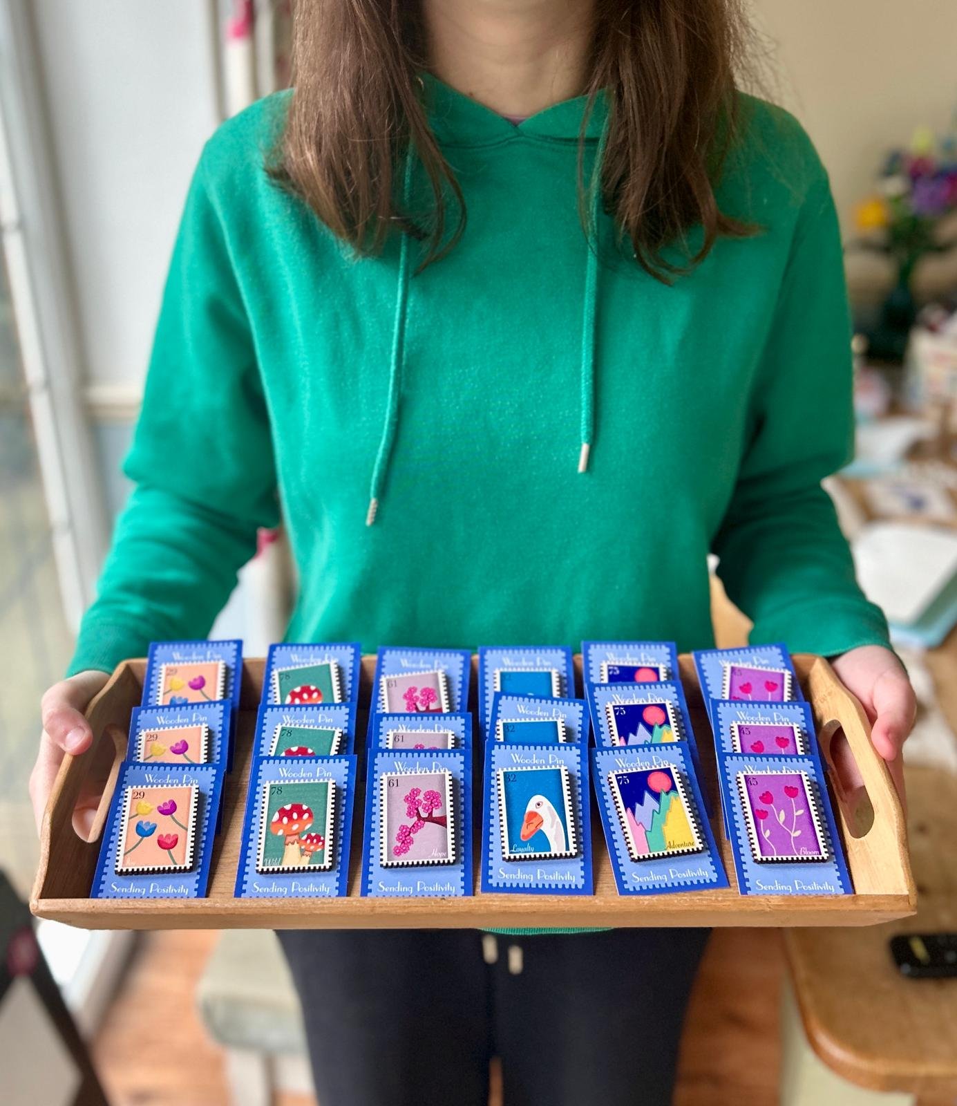 Person wearing a green hoodie holding a wooden tray with colorful painted matchbox covers featuring various nature and animal designs.