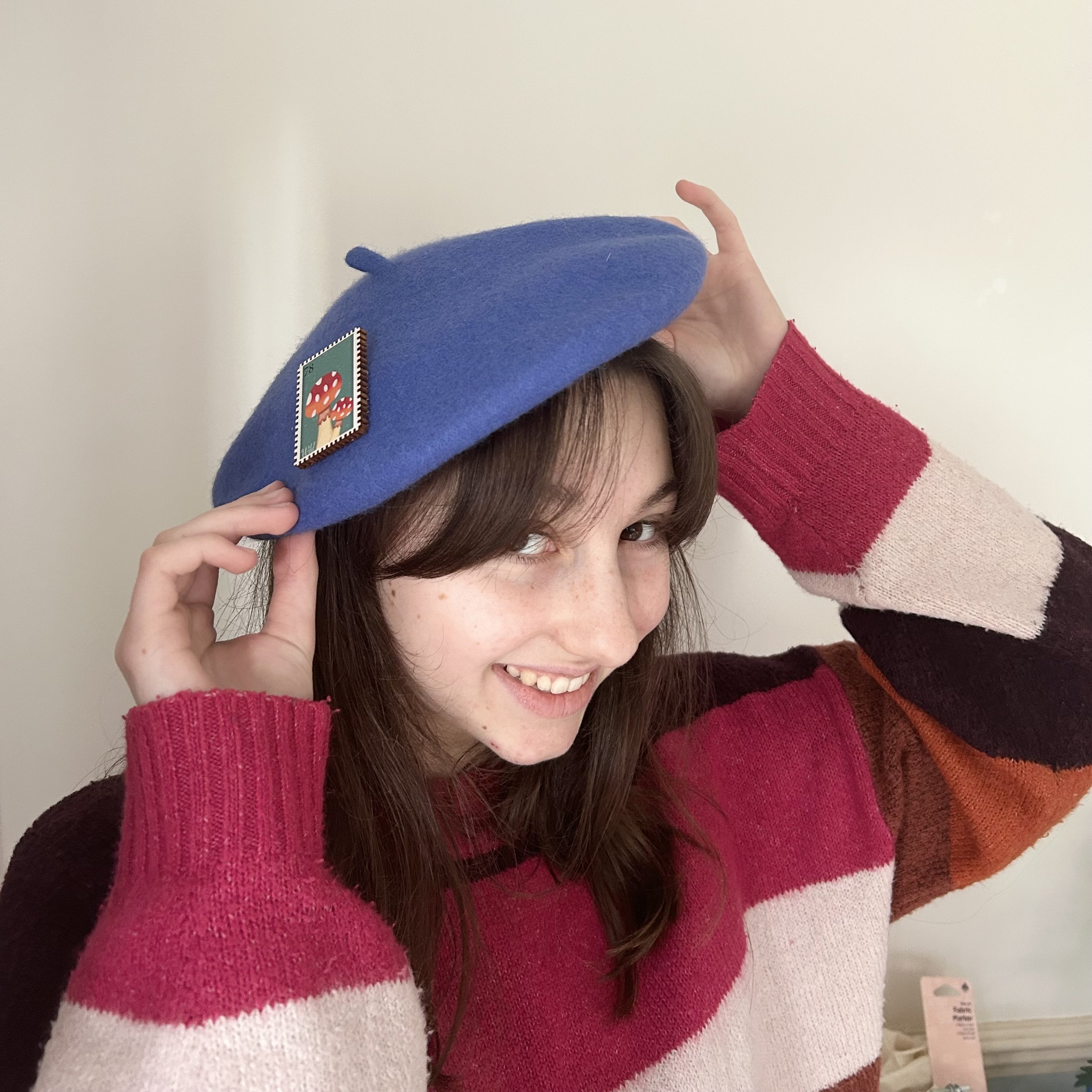 A young woman with brown hair, freckles, and a bright smile, wearing a striped sweater with pink, red, cream, and brown colors, holding a blue beret with a mushroom sticker on it, standing in front of a plain wall.