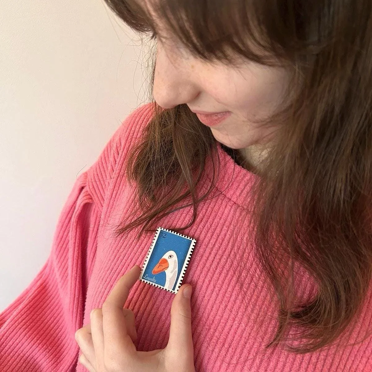 A woman with brown hair wearing a pink sweater, holding a postage stamp featuring a cartoon duck with the word 'Loyalty' on it.