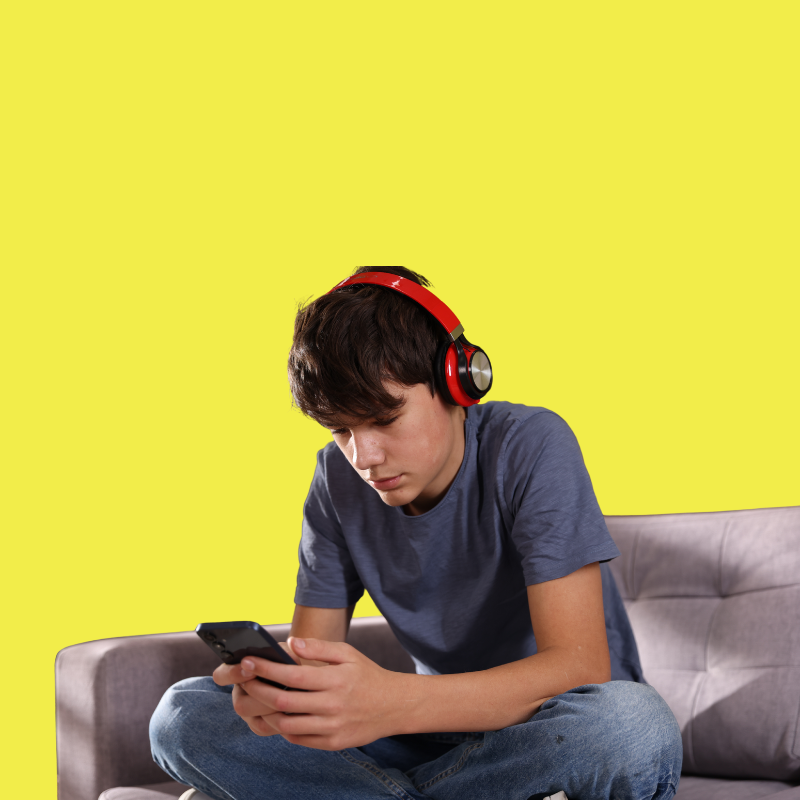 A teenage boy sitting on a gray sofa, wearing a navy blue t-shirt and blue jeans, with red headphones on, looking at a smartphone against a bright yellow background.