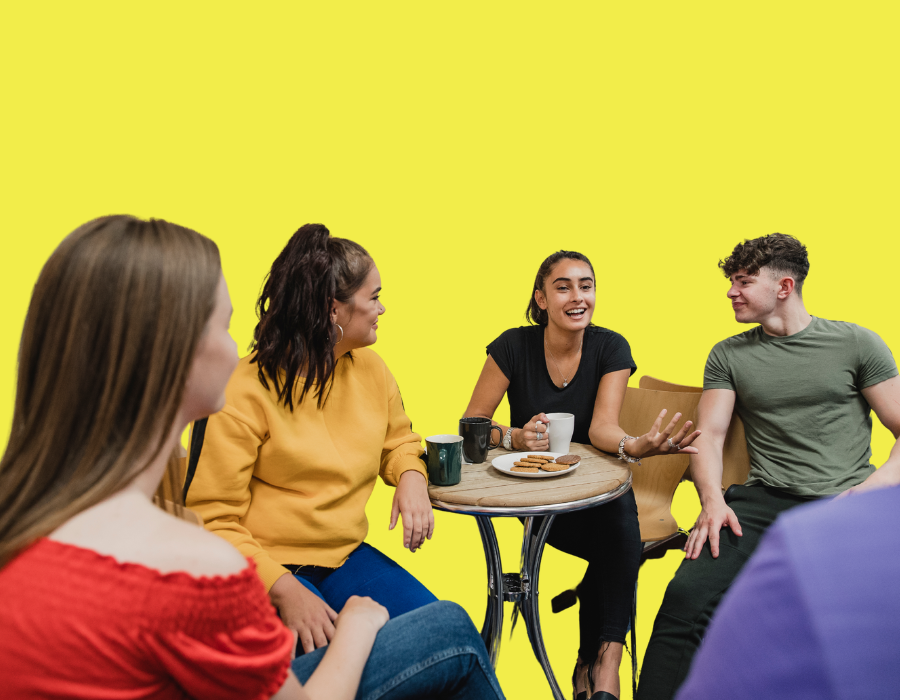 Five young adults sitting around a table, chatting and laughing, with coffee mugs and cookies on the table, against a plain yellow background.