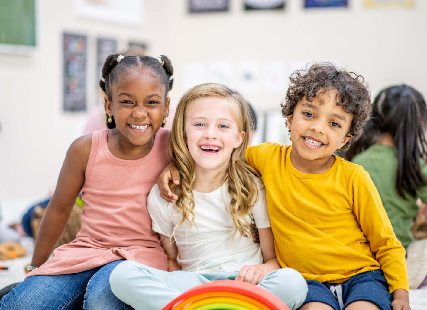 Three children in a classroom taking a picture. They are sitting criuss cross and all smiling at the camera.