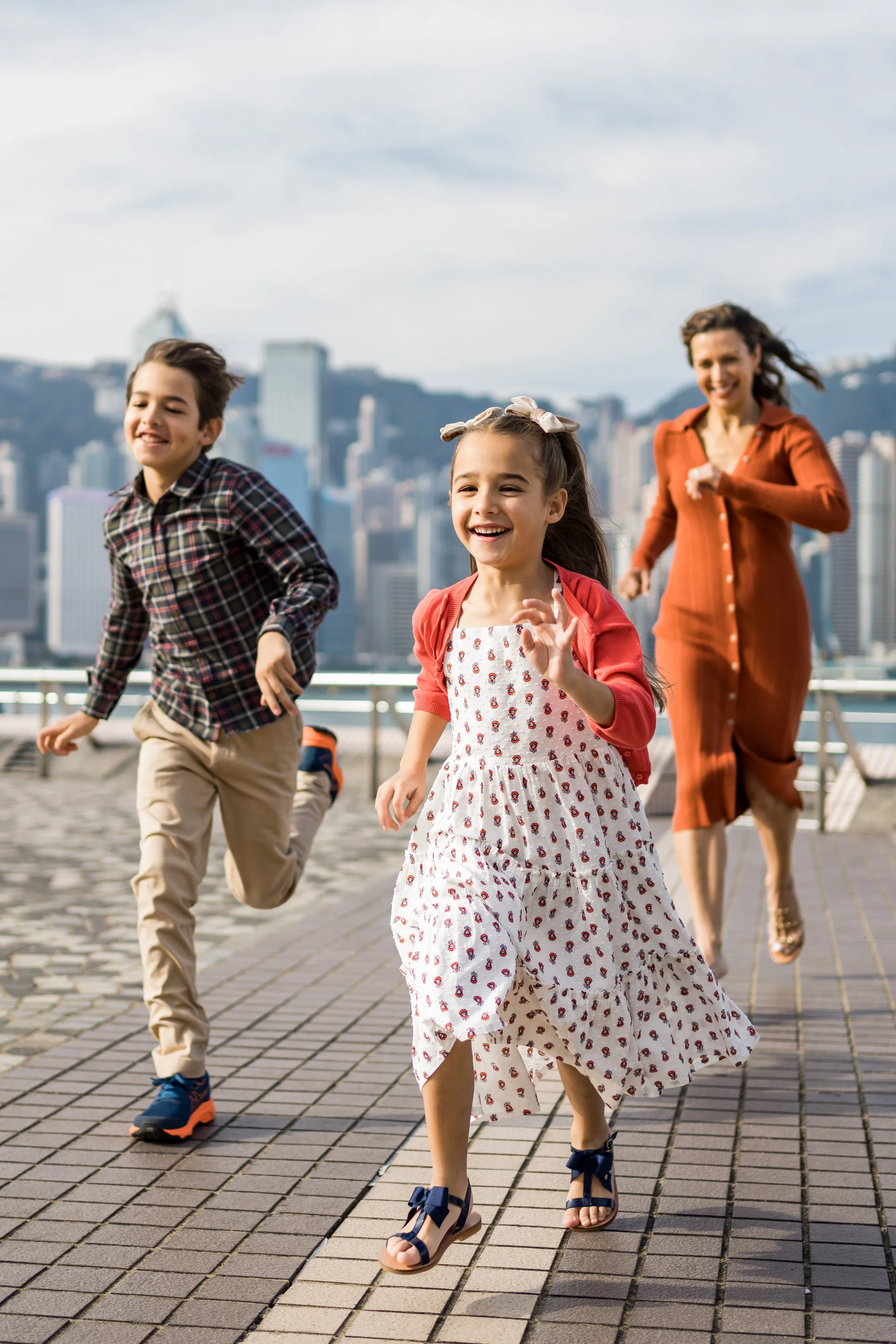 A picture of two children laughing and running and their mother running behind them. They are by a  harbor, with a city skyline behind them.