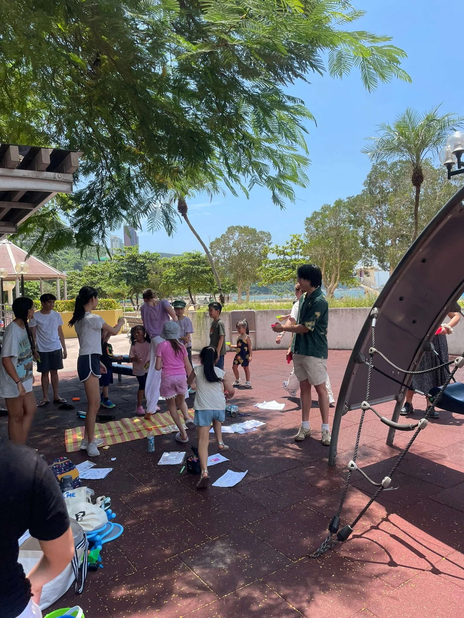 A group of children and adults gathered under a shaded area at a playground while engaging in a group activity on a sunny day. There are trees, a hammock, and playground equipment visible.