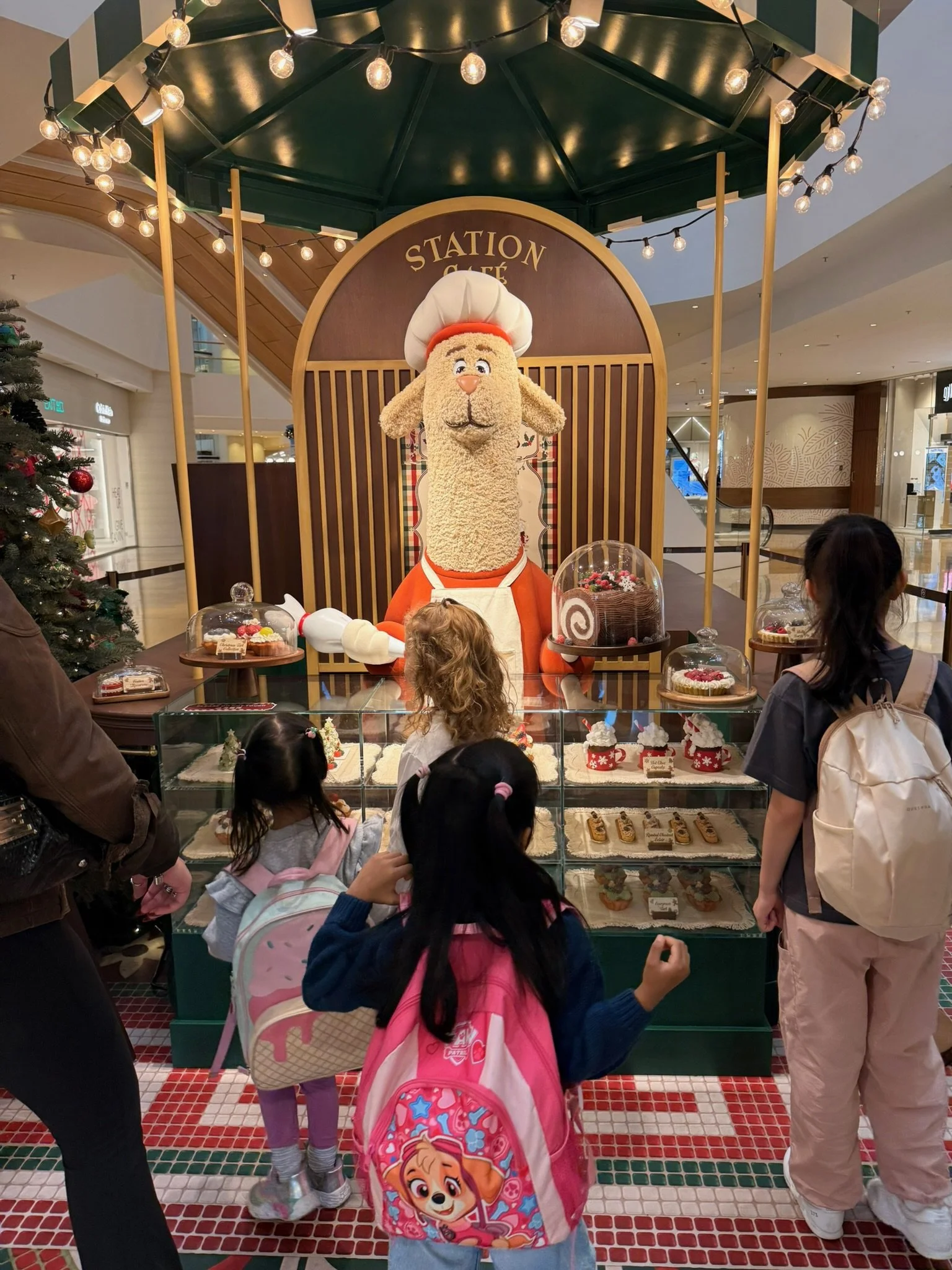 Children and adults looking at a display of Christmas desserts featuring a large puppet of a dog dressed as a chef, with the sign 'Station Café' above it, inside a shopping mall.