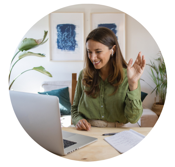 A therapist is on an online meeting and waving at her computer. She is seated in her office.