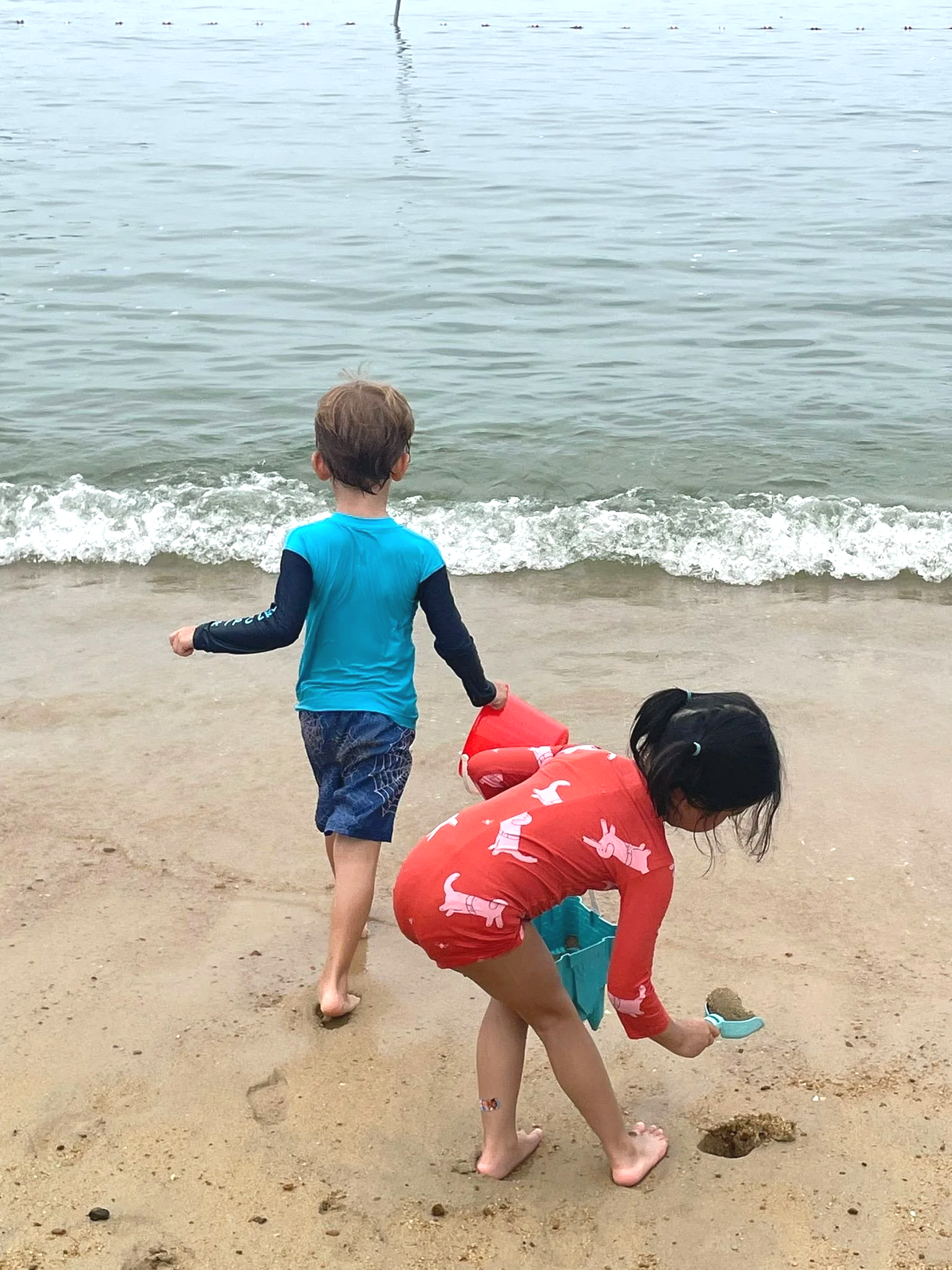 Two young children playing at the beach. One is running toward the ocean with a bucket in his hand. The other one is filling a sand castle mold with a toy shovel.