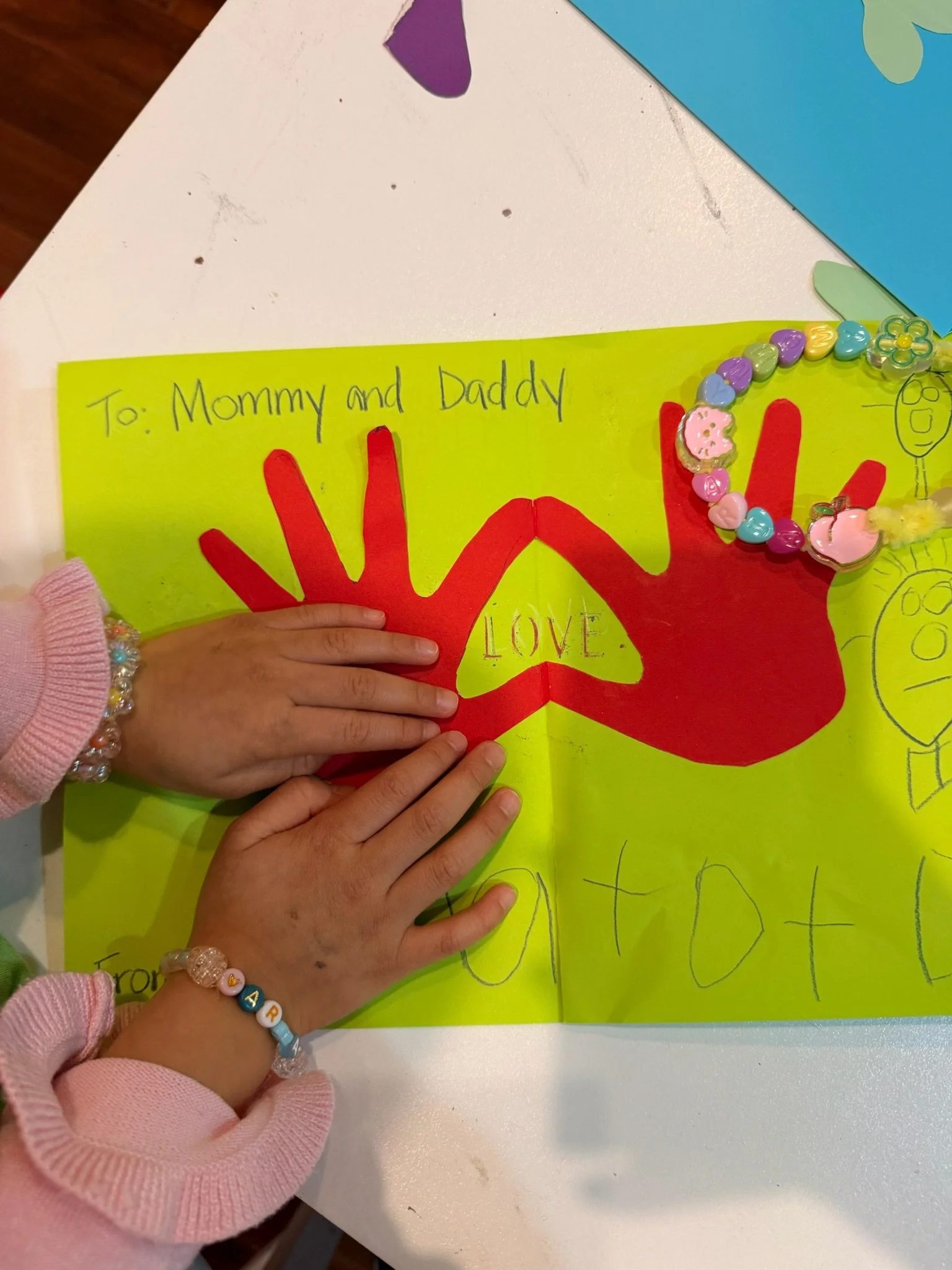 Children's hands with bracelets place on a bright yellow poster with red handprints and the words "LOVE" and "happy" written on it, and a handwritten note at the top reads "To: Mommy and Daddy."