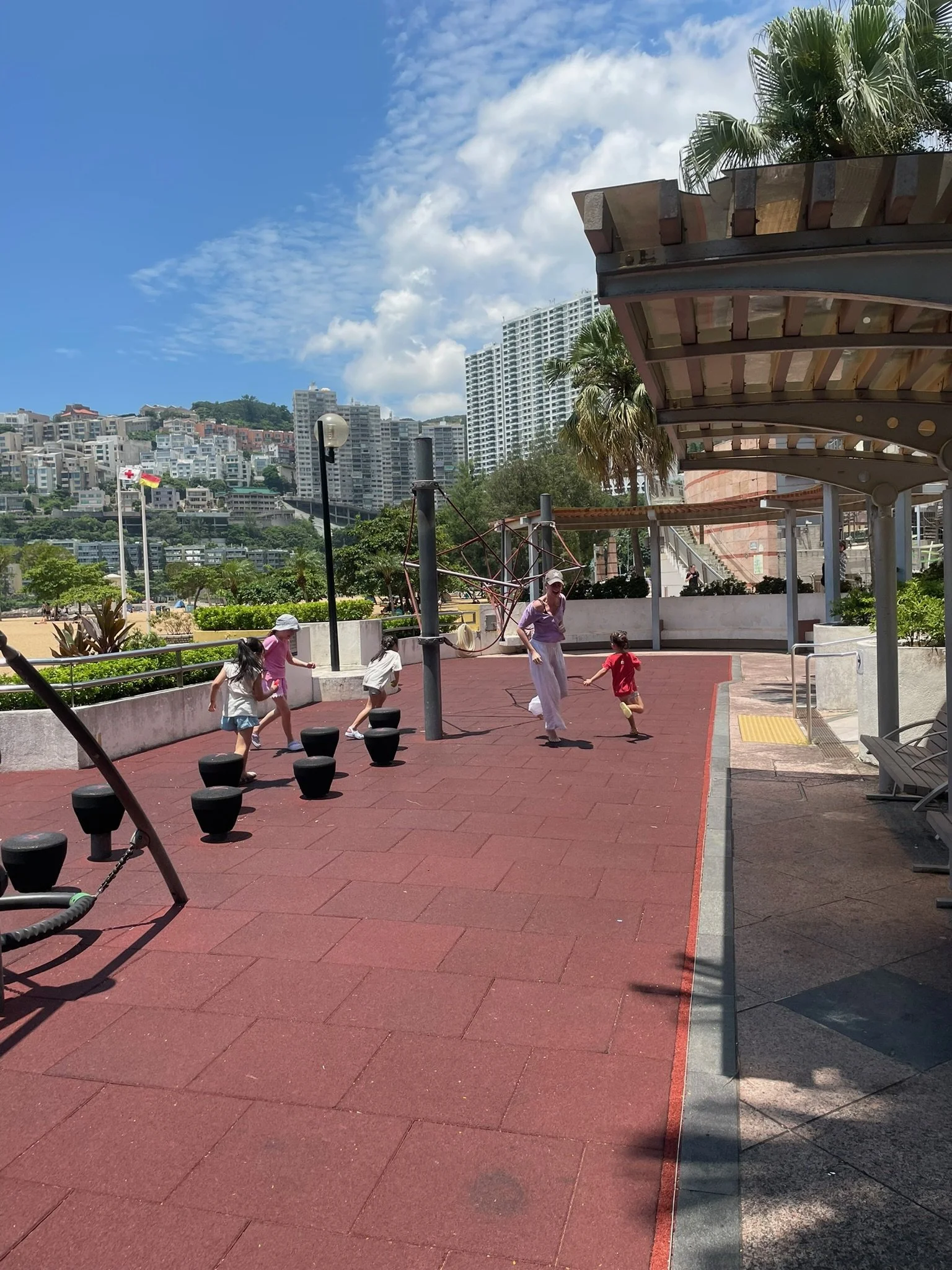 Children and adults playing at an outdoor playground on a sunny day, with a cityscape of high-rise buildings and hills in the background, and palm trees around.