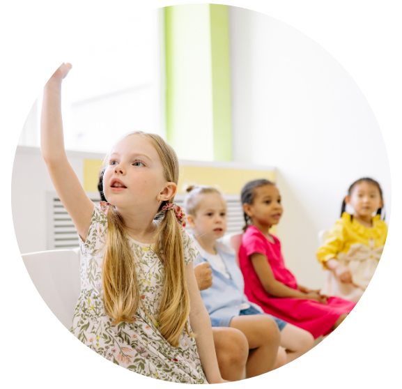 A younge girl is raising her hand and looking expectantly towards the front of the classroom.