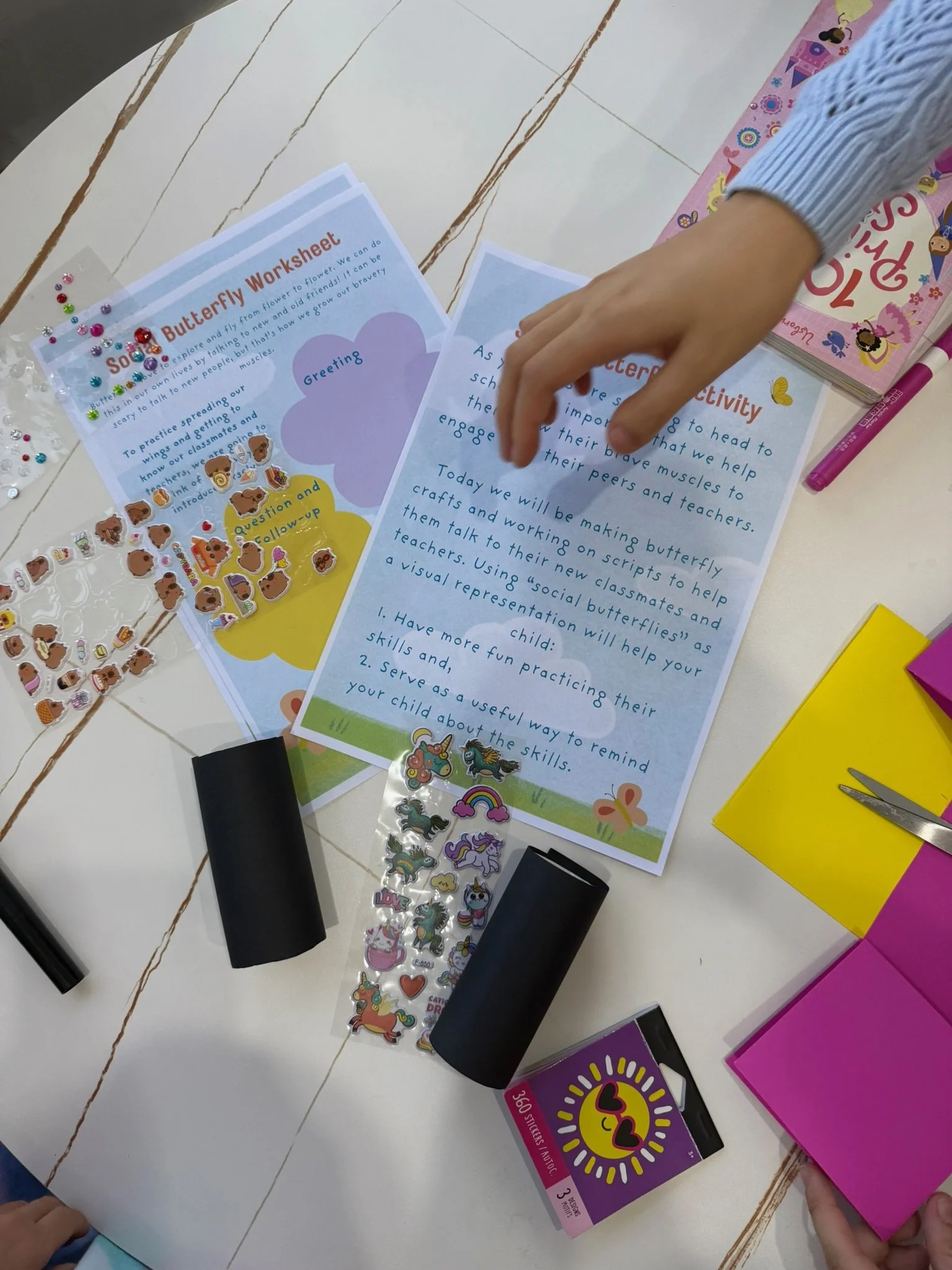 A table with papers, books, stickers, and coloring supplies. A child's hand is reaching to pick up a sheet of paper with text about a butterfly activity.