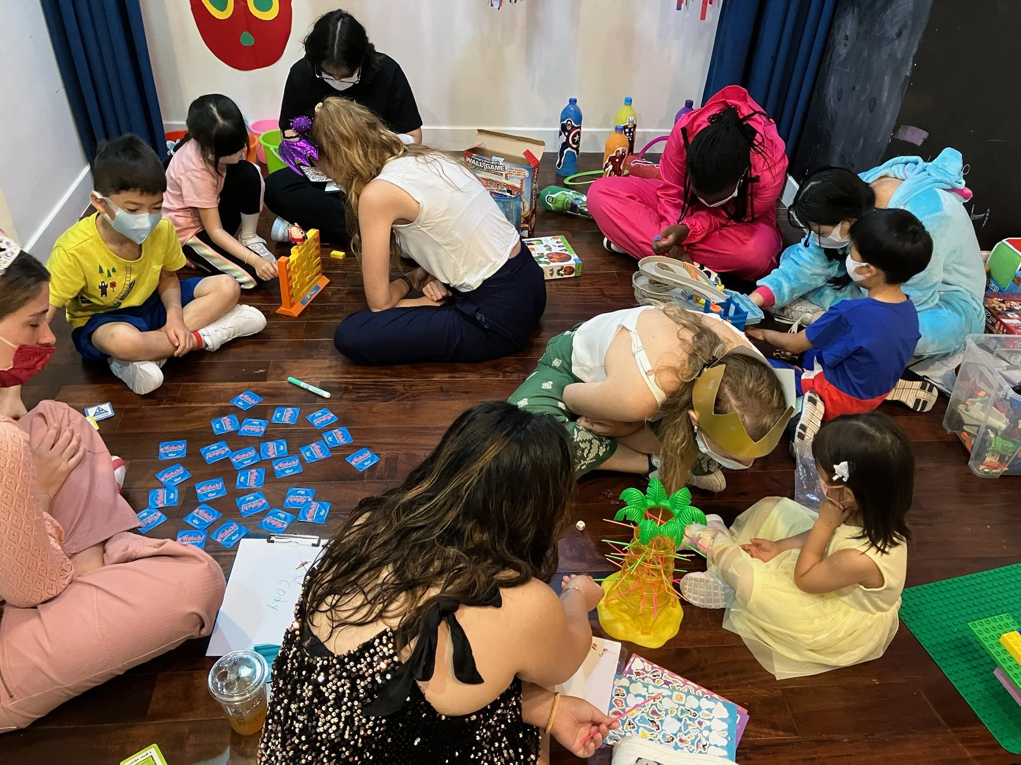 Children and adults sitting on a wooden floor playing board games and building with toys, with some wearing face masks.