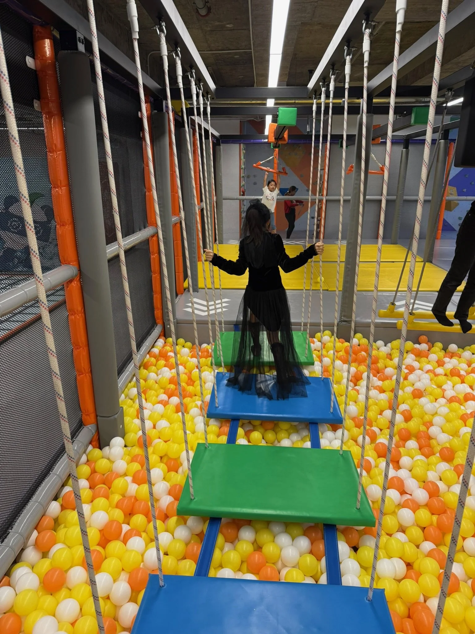 Children playing on a stairway with colorful platforms and ropes in an indoor playground, with a ball pit filled with yellow, white, and orange balls at the bottom.