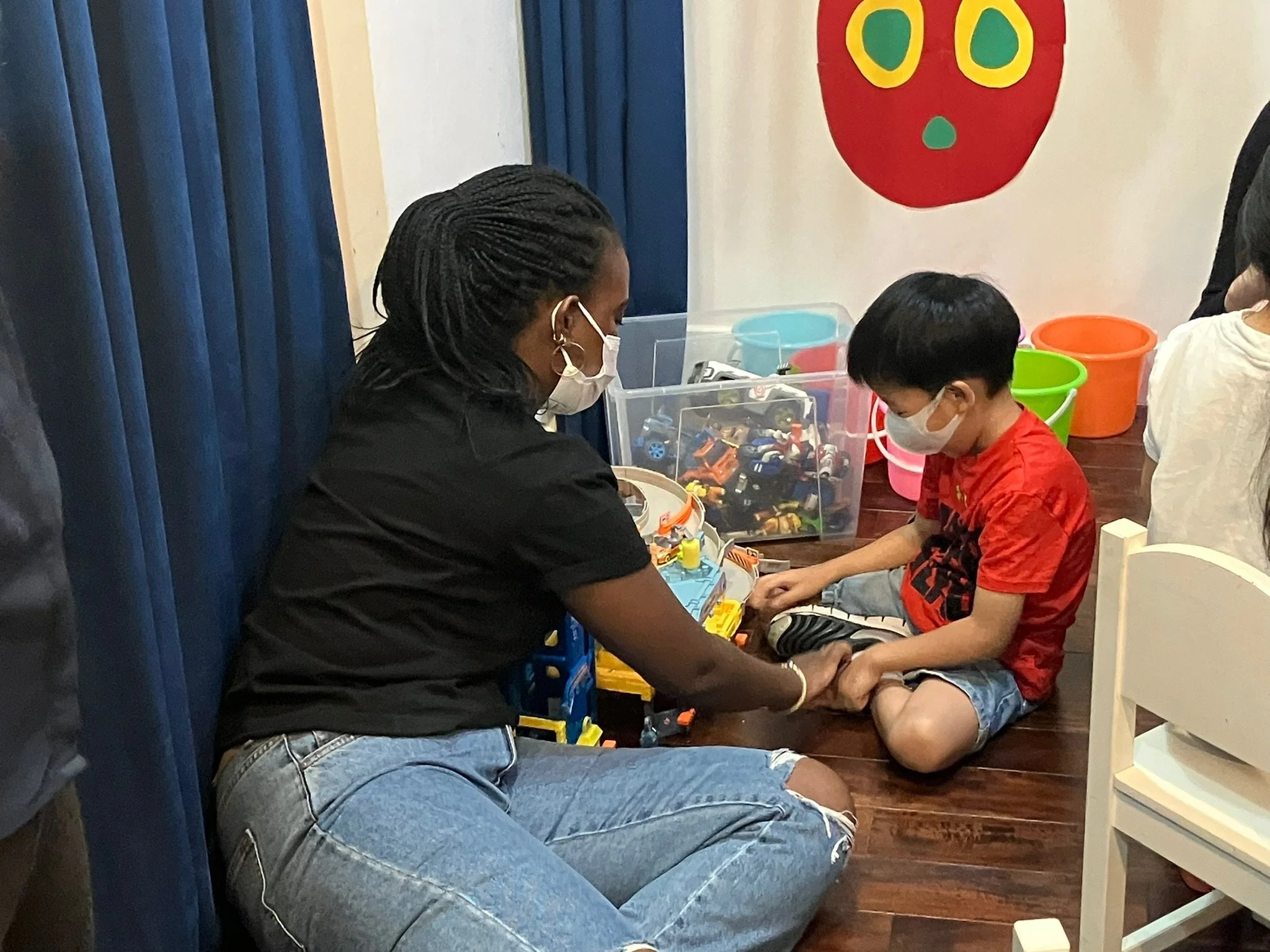 A woman and a young boy wearing face masks playing with toy cars on the floor.