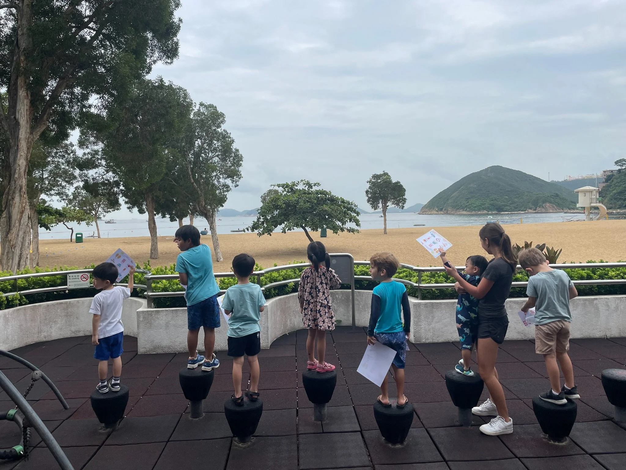 Children and a woman standing on balance pods at a park overlooking a beach, trees, water, and islands with cloudy sky.