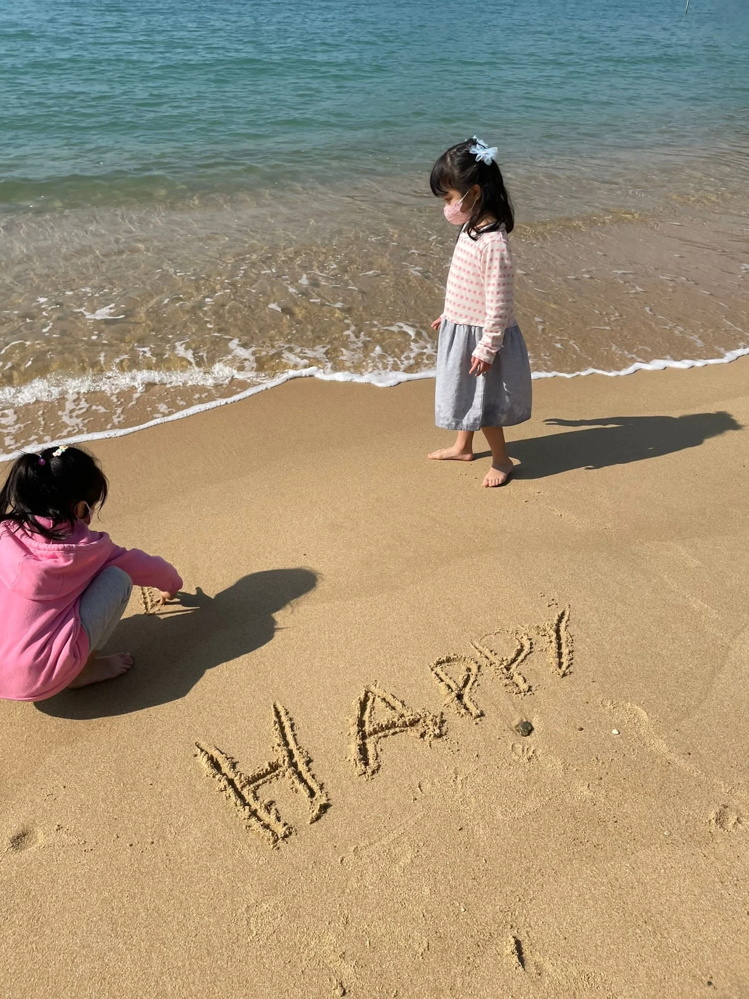 Two young girls are on a sandy beach with the ocean in the background. One girl is squatting and writing 'HAPPY' in the sand, while the other girl stands nearby, looking at the sand. Both girls are wearing masks and casual clothing.