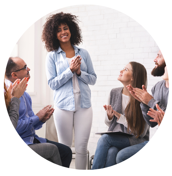 A woman is standing while adults clap for her. The adults are seated in a circle in chairs.