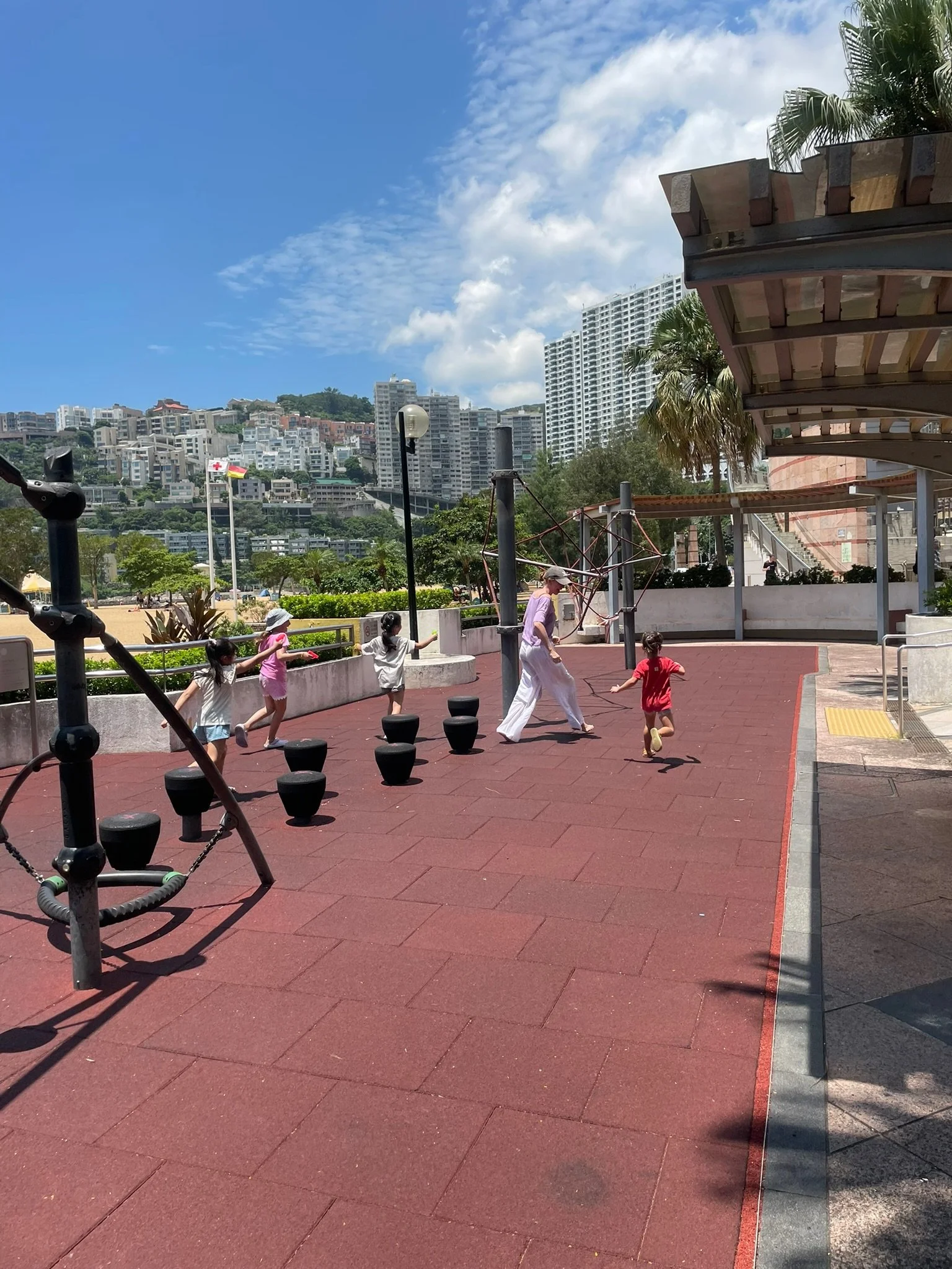 Children playing on a playground with an adult supervising on a sunny day in a park with city buildings in the background.