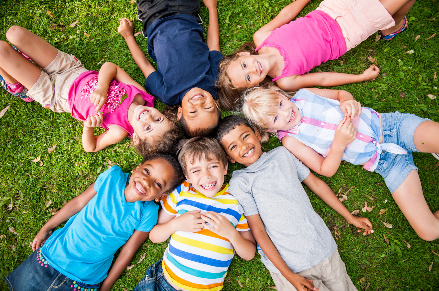 A picture of seven children lying on their backs on green grass. They are in a circle formation and smiling up at the camera.