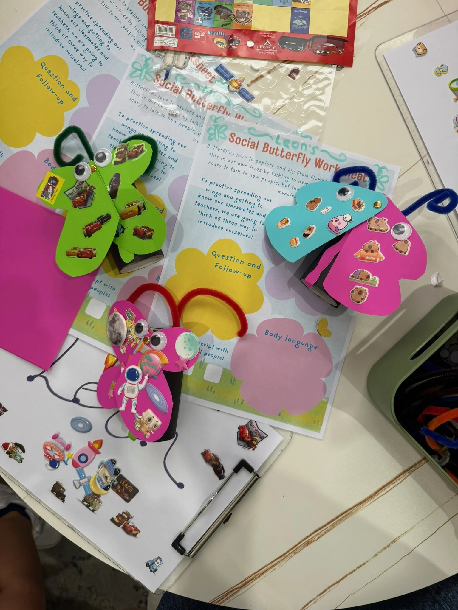 Colorful paper crafts of butterflies with googly eyes, stickers, and pipe cleaners on a desk with educational materials about social butterfly behavior and body language.