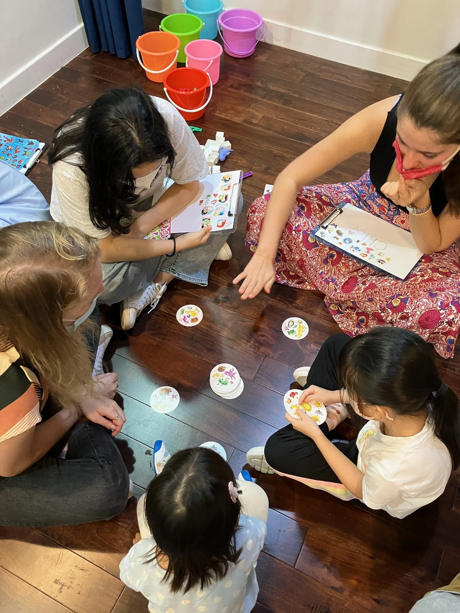 A group of children and a woman sitting on a wooden floor playing a sticker or card game with colorful round cards. There are buckets of different colors in the background.