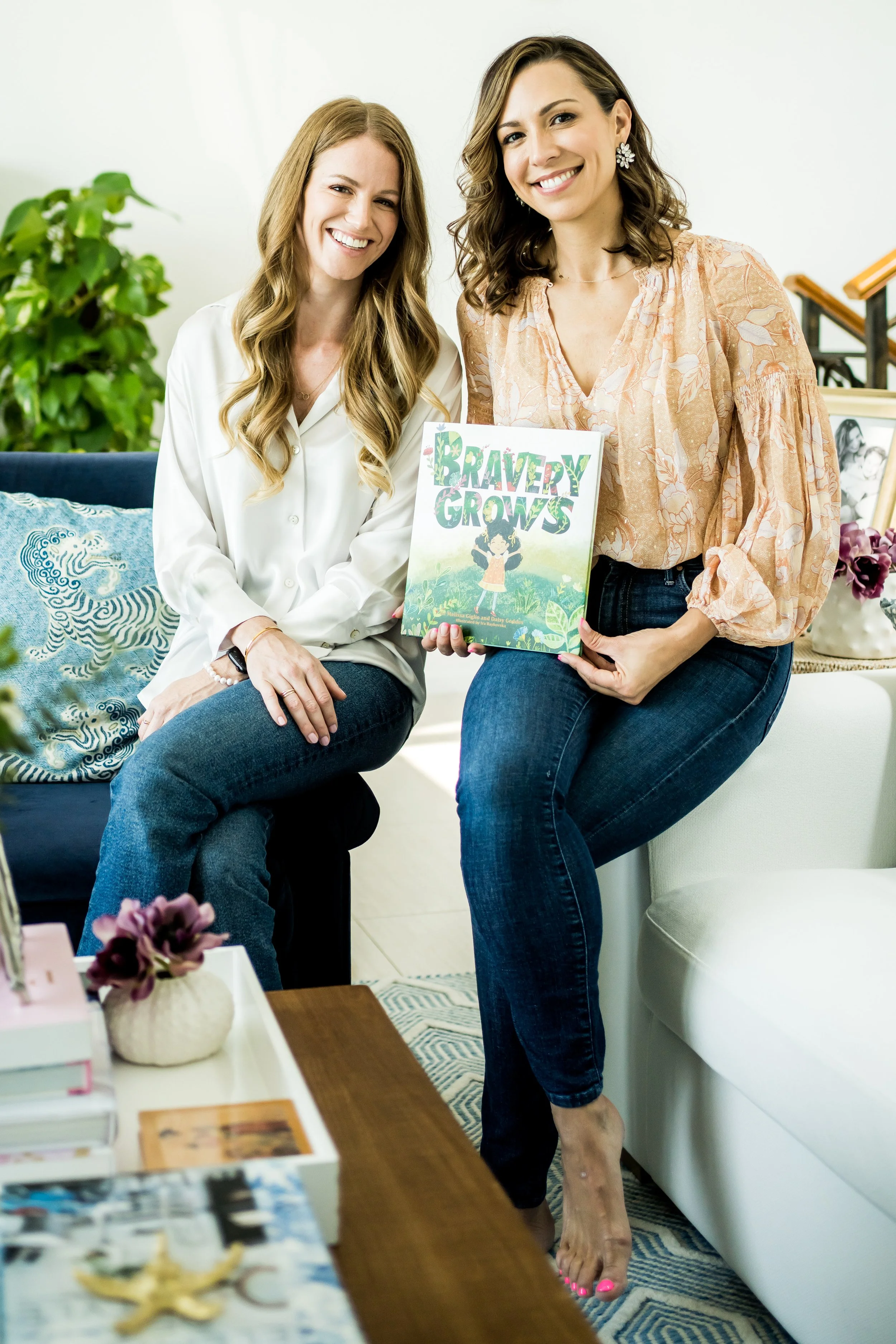 Dr Melissa Giglio and Daisy Geddes smilingat and holding their book "Bravery Grows". They are sitting on the edge of a two different couches, next to each other.